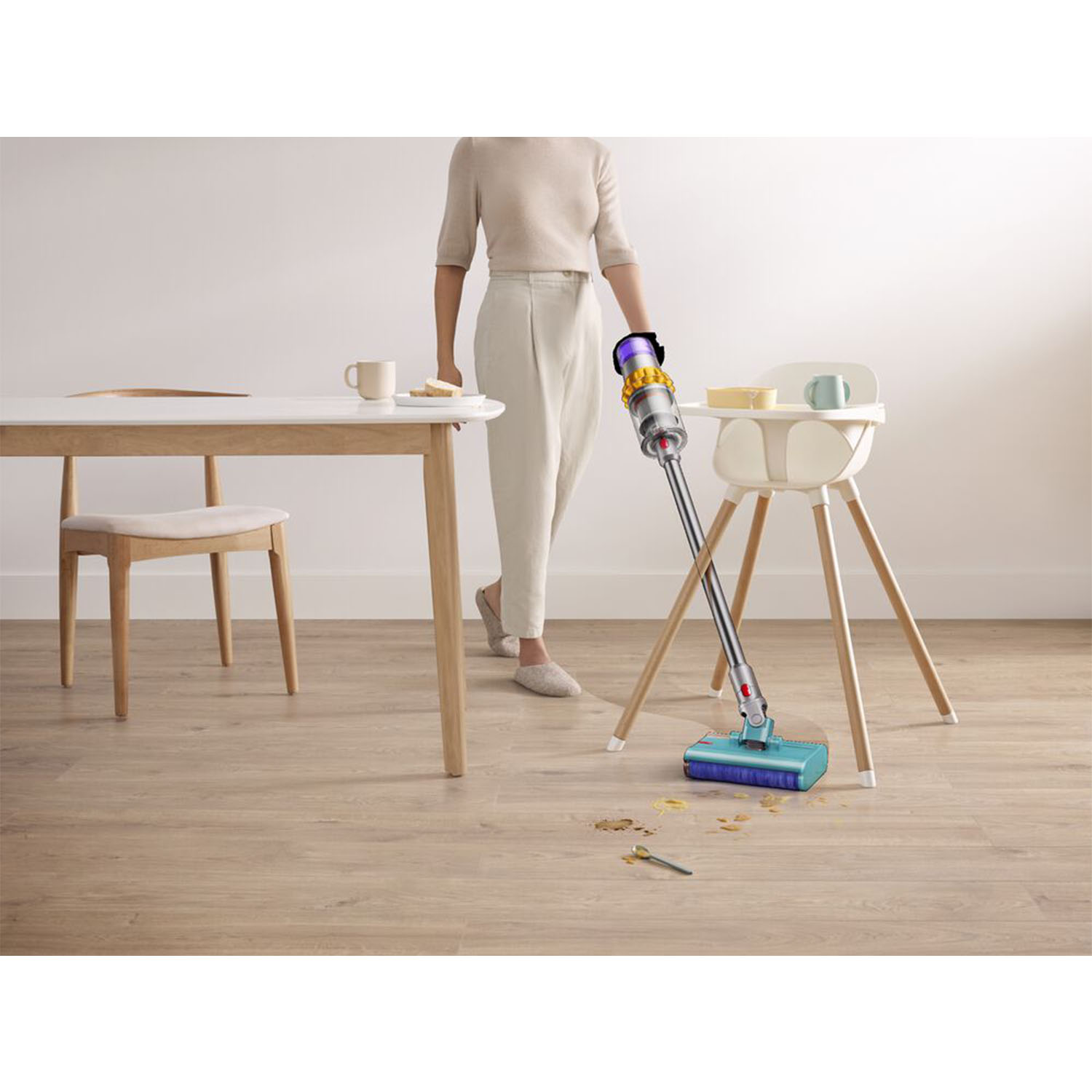 Una donna passa l'aspirapolvere sul pavimento con un Dyson. Vicino, un seggiolone e un tavolo con cibo.