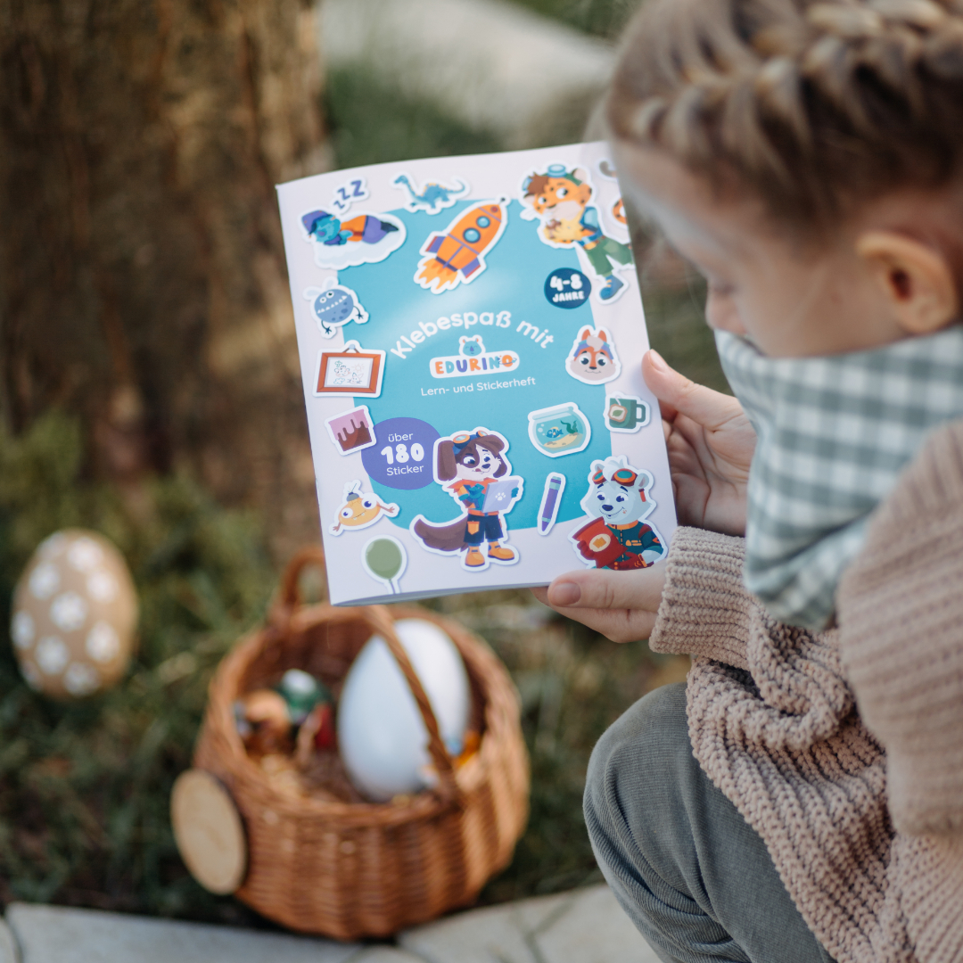 Ein Mädchen betrachtet ein Stickerbuch mit dem Titel 'Klebespaß mit Edurino'. Es gibt ein Ei und einen Korb.