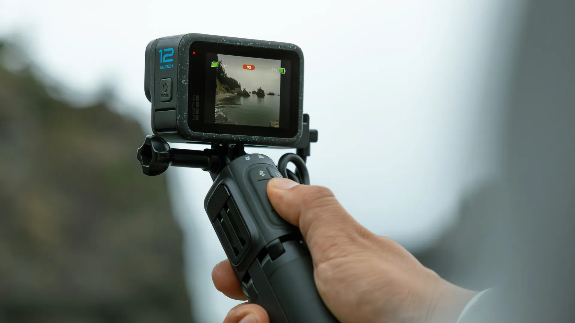 Egy személy fekete GoPro kamerát tart egy szelfiboton, egy kültéri jelenetet rögzítve.