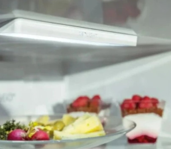 Wnętrze lodówki z produktami spożywczymi na szklanych półkach. Zawiera ser, pomidory, ogórki kiszone i desery w kubkach.