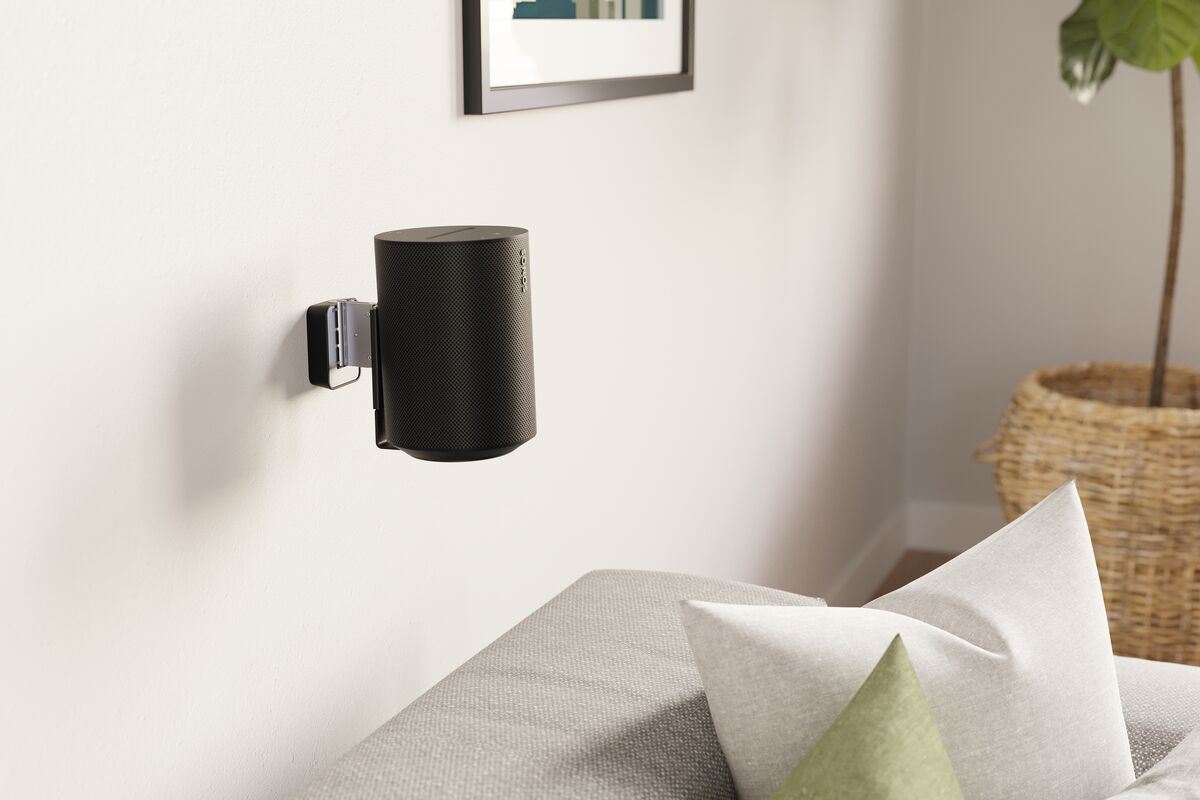 Un altavoz negro montado en una pared blanca con un sofá gris y una planta en el fondo.