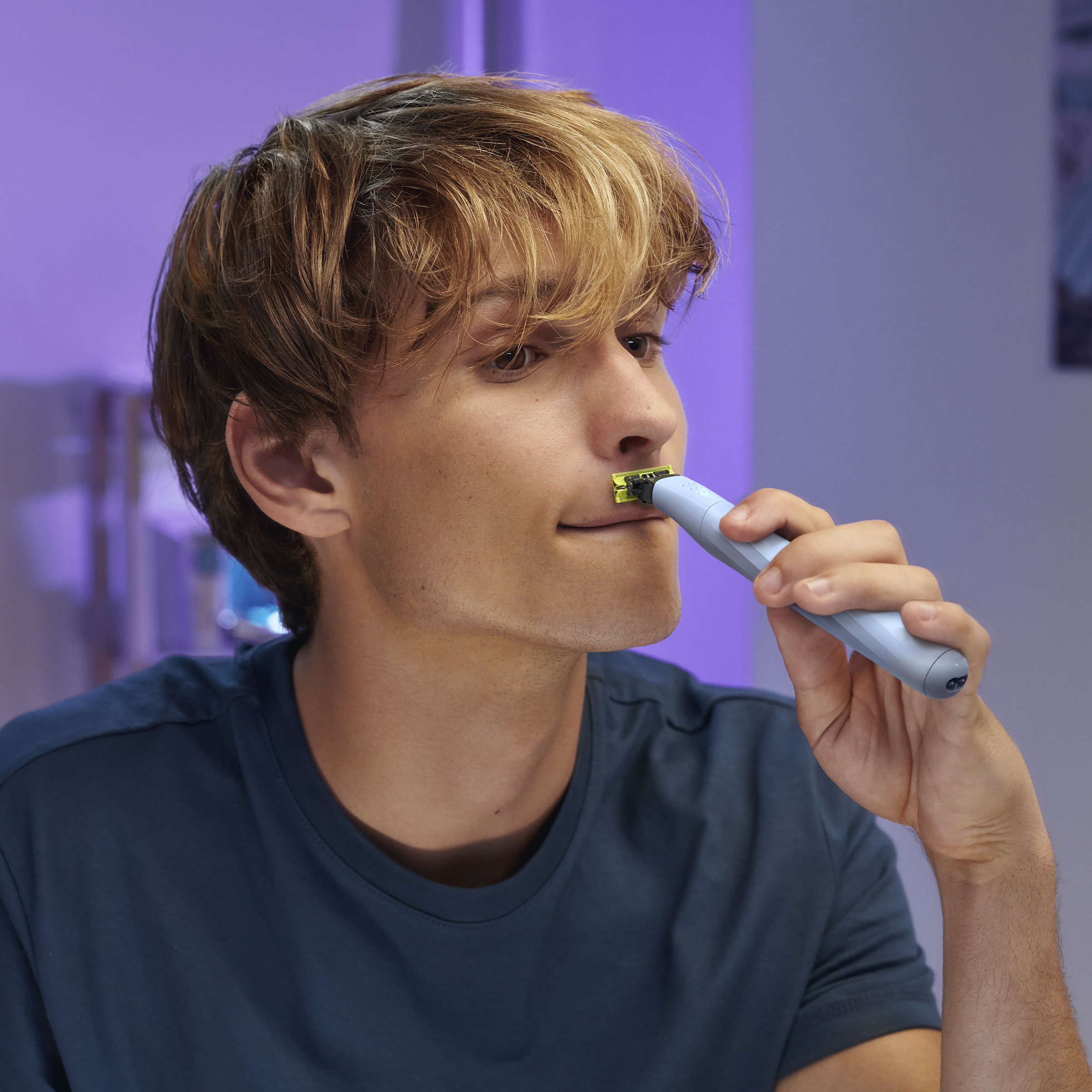 Mężczyzna goli twarz niebiesko-żółtą maszynką. Znajduje się w łazience z fioletowym oświetleniem.