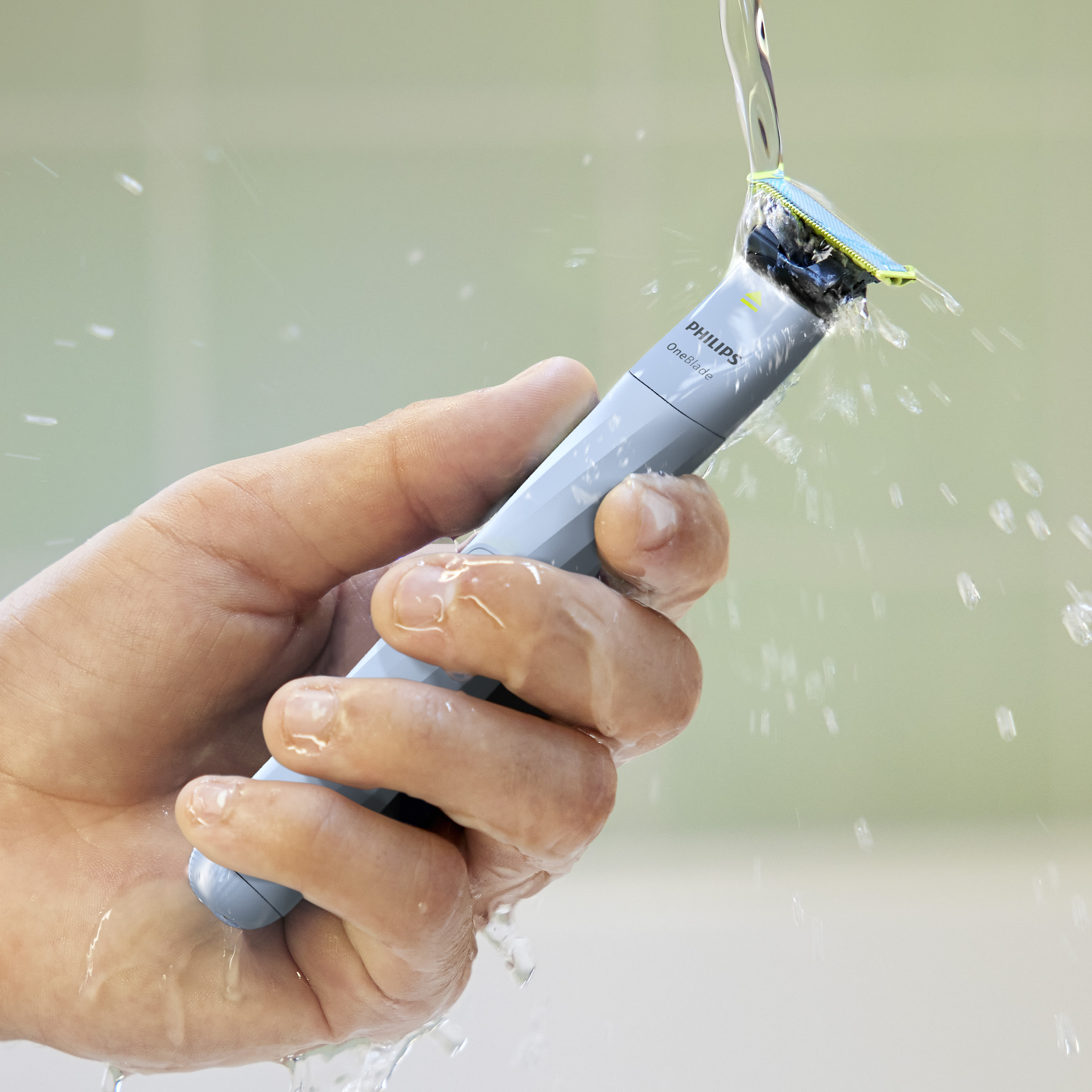 Dłoń trzyma golarkę Philips OneBlade pod bieżącą wodą. Golarka jest szara i niebieska.