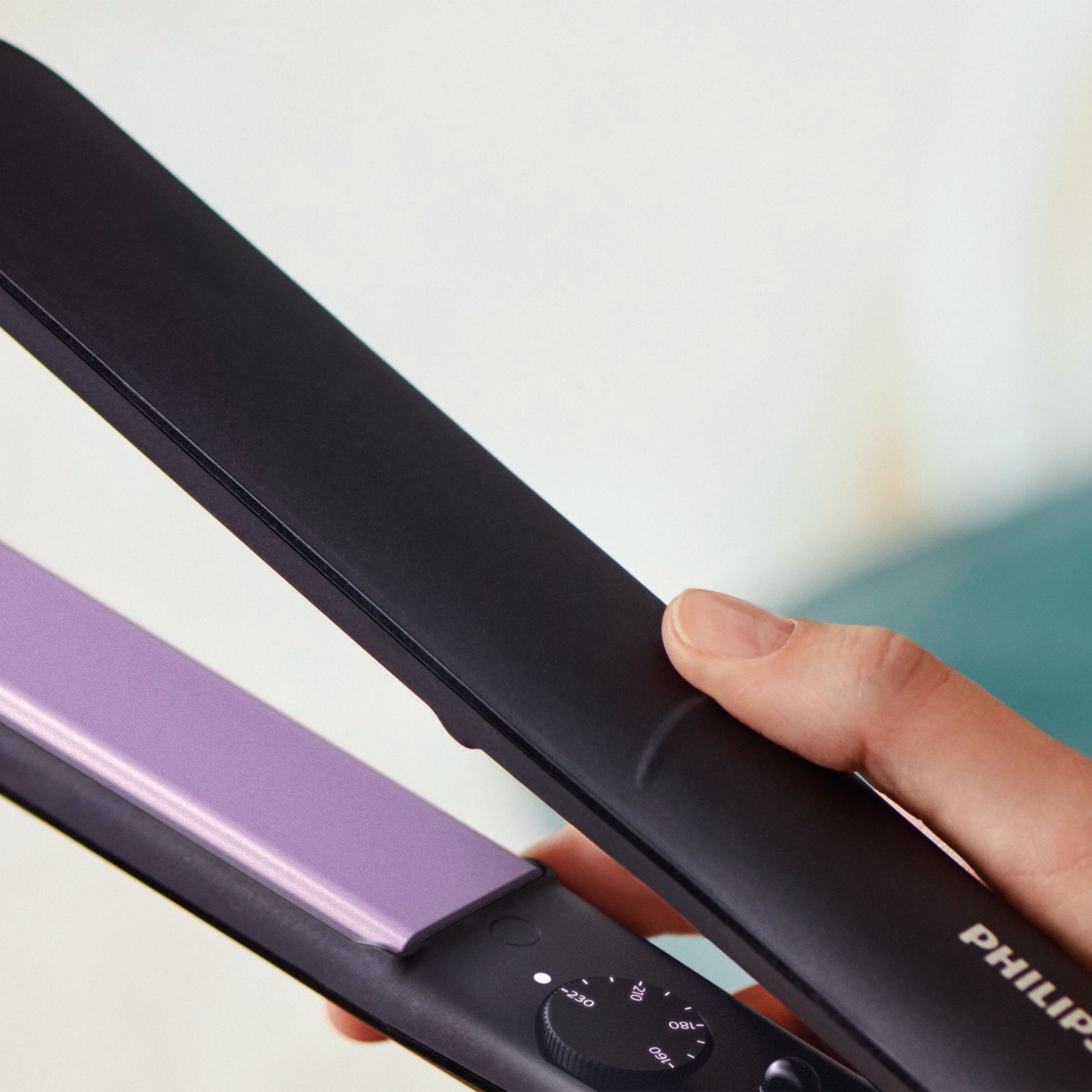 Czarna prostownica do włosów z fioletową płytką. Dłoń ją trzyma, rozmyte tło.