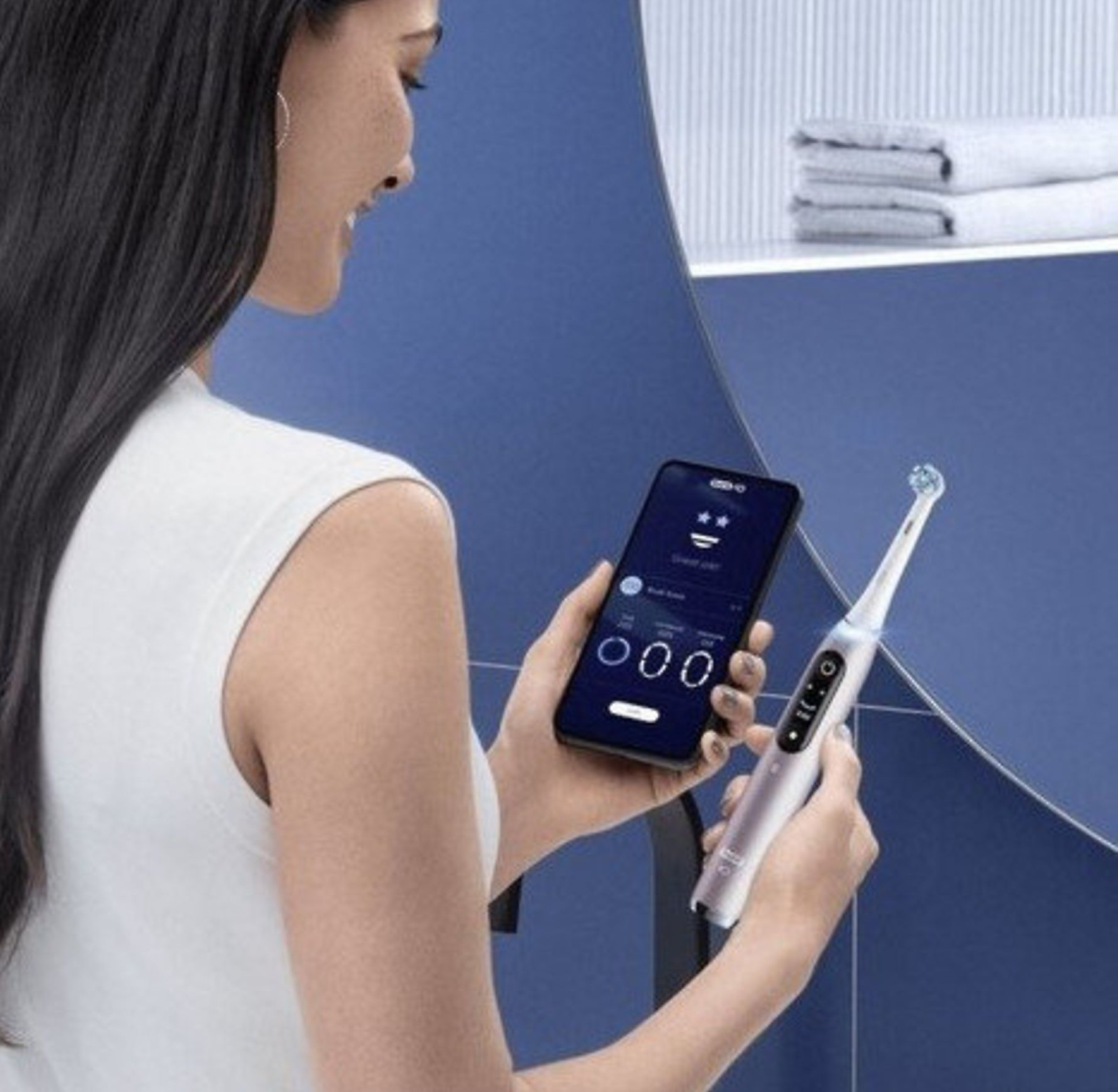 Kobieta trzymająca smartfon i białą elektryczną szczoteczkę do zębów przed lustrem w niebieskiej łazience.