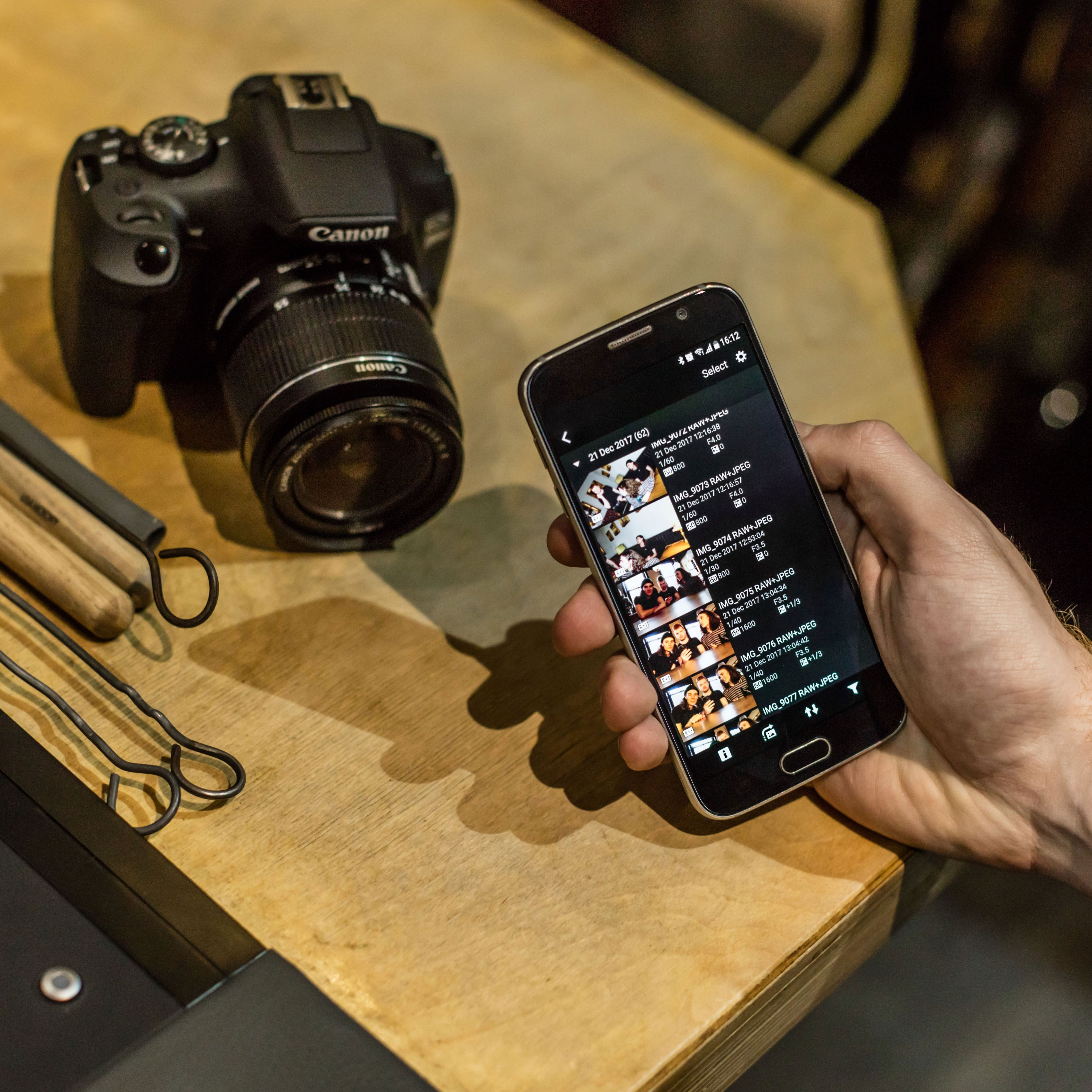 Persona che tiene un telefono con foto, accanto a una fotocamera Canon su un tavolo di legno.