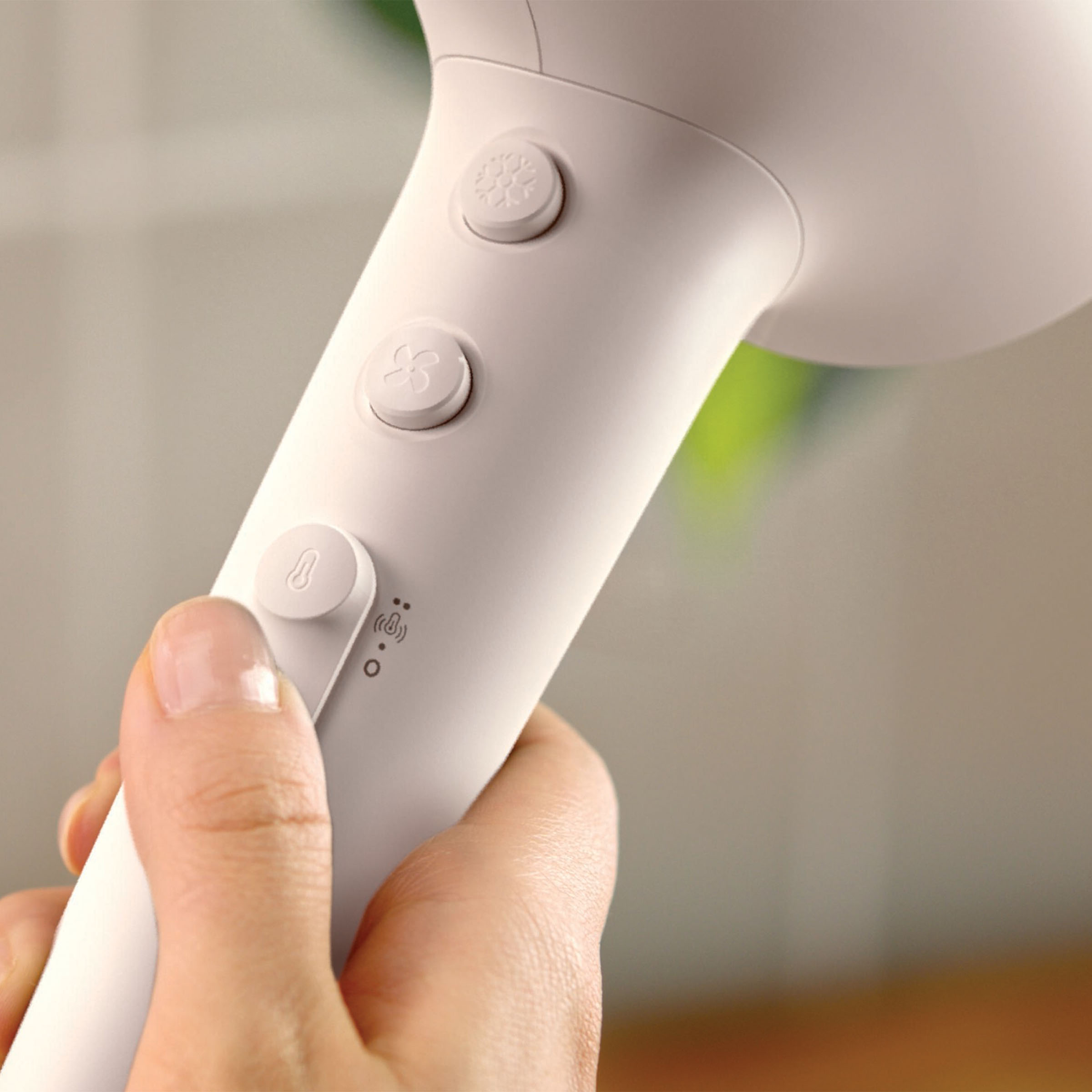 Zbliżenie dłoni trzymającej białą suszarkę do włosów. Widoczne są przyciski przepływu powietrza i temperatury.