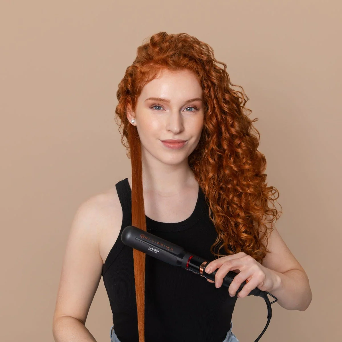 Femme aux cheveux roux bouclés utilisant un fer à lisser. Elle porte un débardeur noir, souriante. Fond beige.