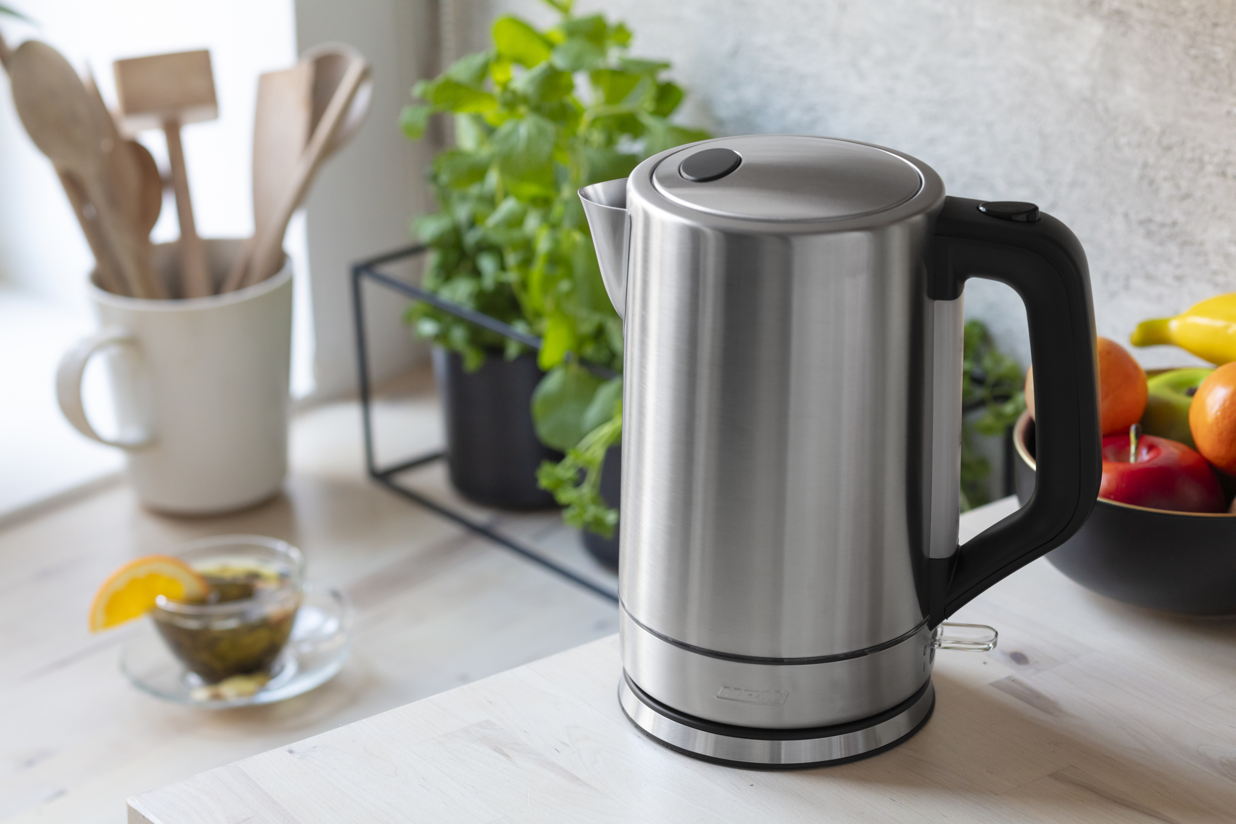 Czajnik elektryczny ze stali nierdzewnej na blacie. W tle filiżanka herbaty, owoce i rośliny.