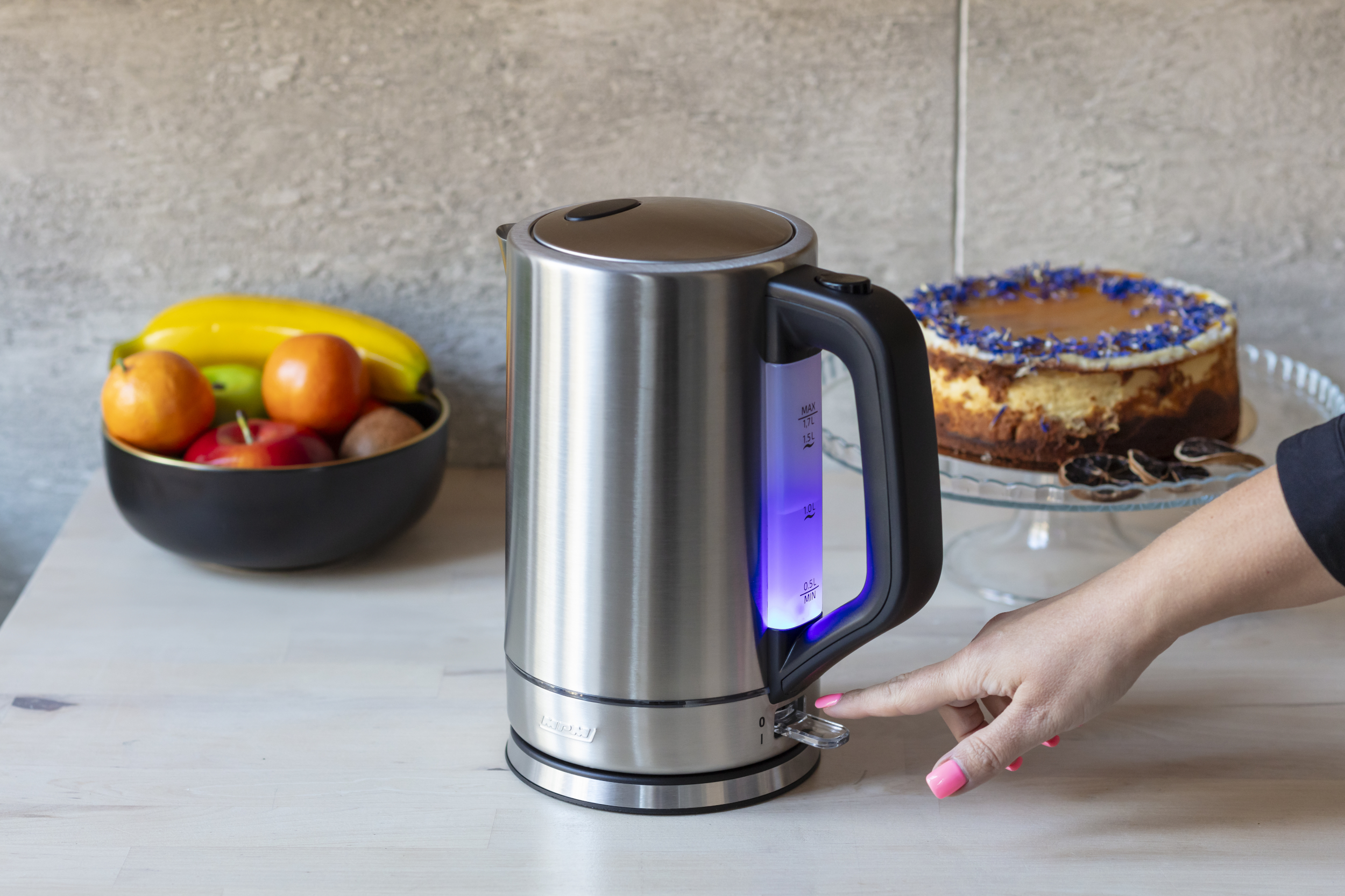 Srebrny czajnik elektryczny, ręka naciska przycisk, z miską owoców i ciastem. Wnętrze.