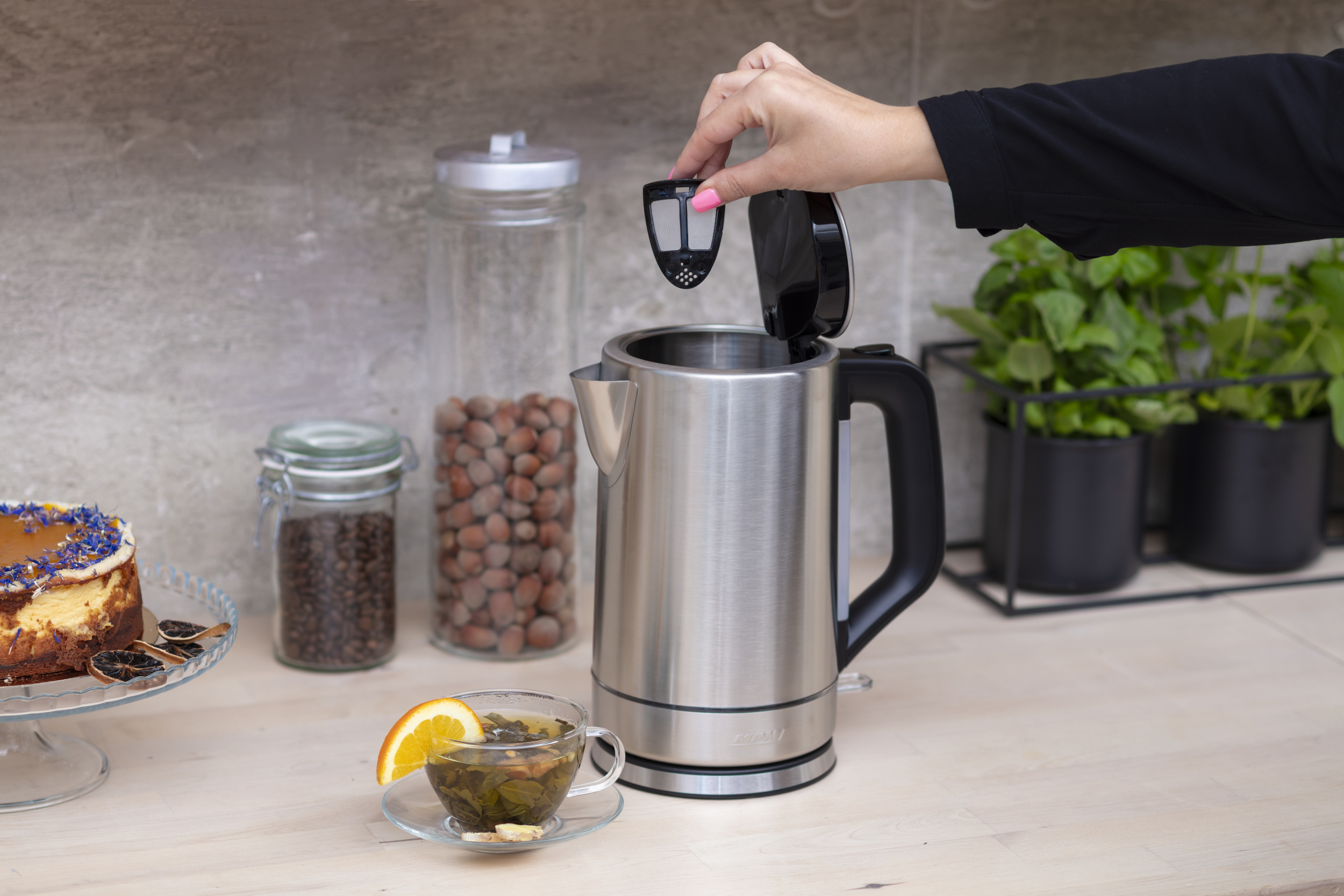 Osoba dodająca zaparzacz do herbaty do czajnika ze stali nierdzewnej. Na blacie są inne przedmioty.