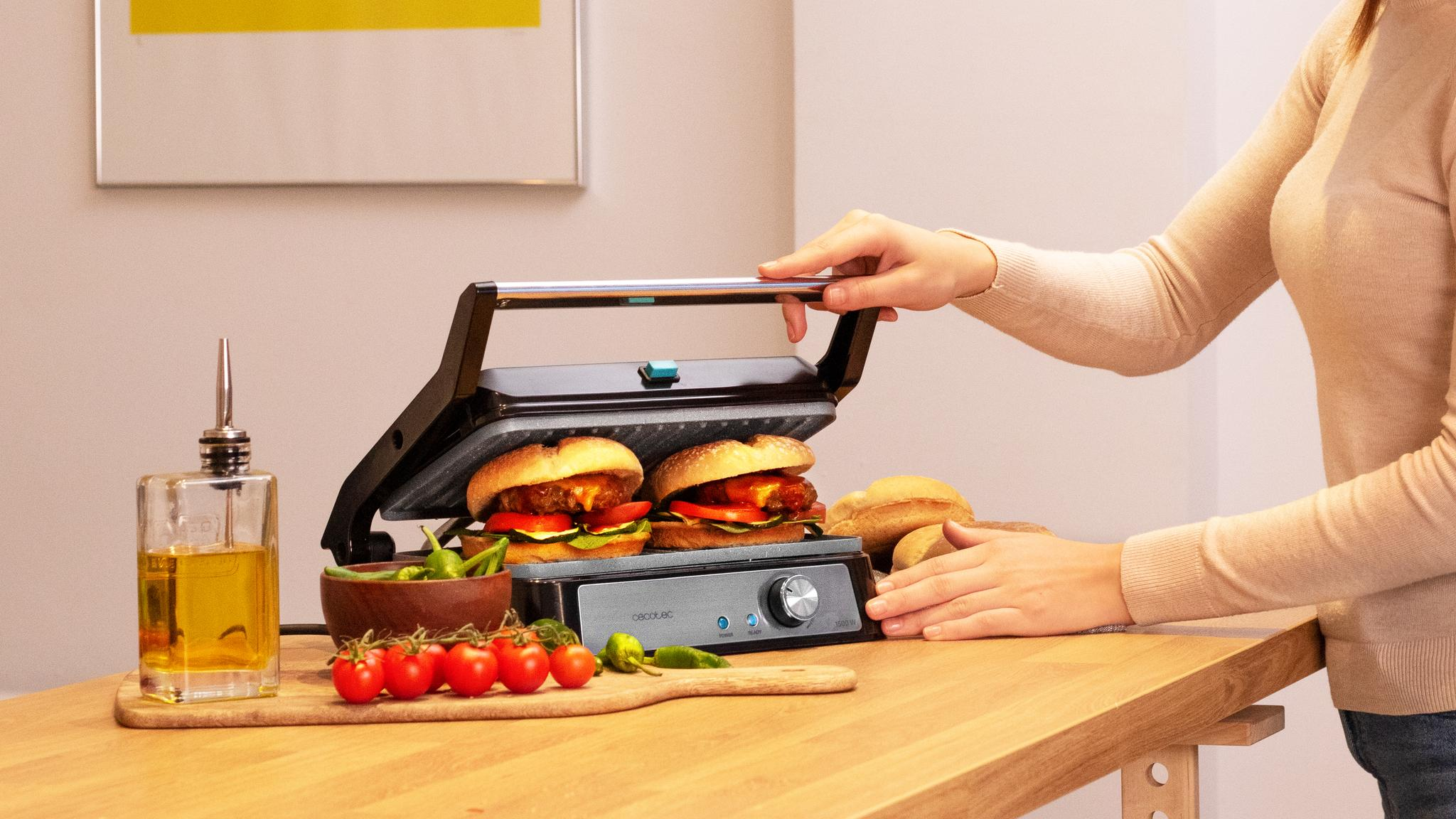 Una mujer usa una parrilla con dos hamburguesas. Tomates, aceite y pan sobre la superficie de madera.