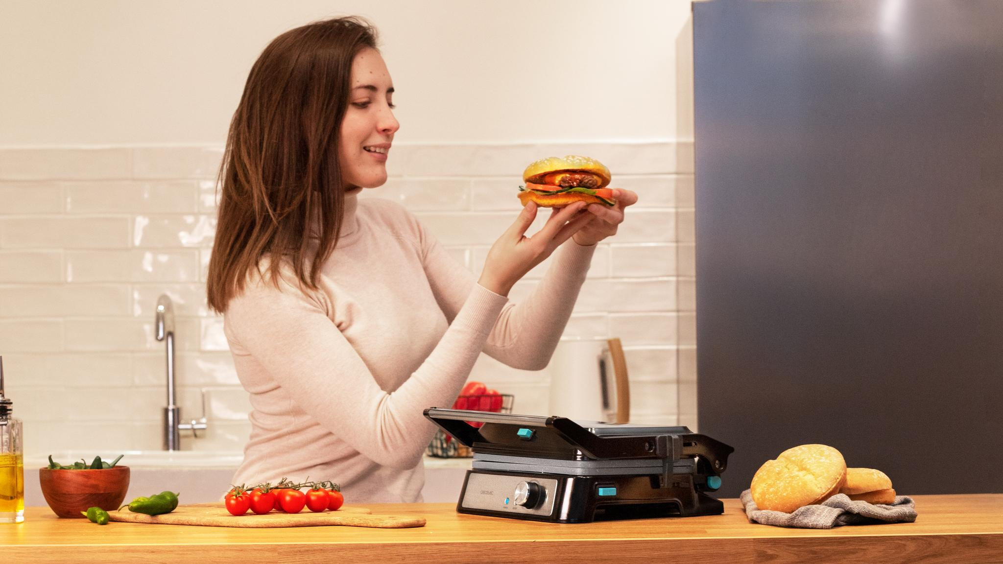 Una mujer sosteniendo una hamburguesa, con una parrilla e ingredientes en una encimera.