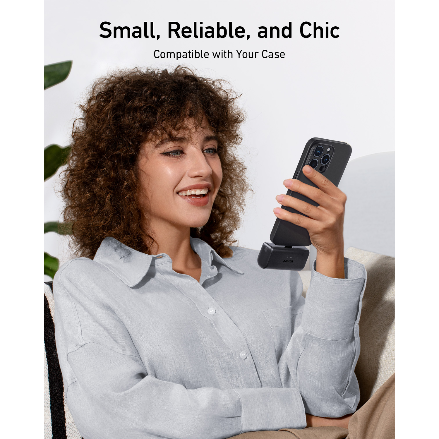 A woman holds a phone with a black case and a charger attached. She smiles.