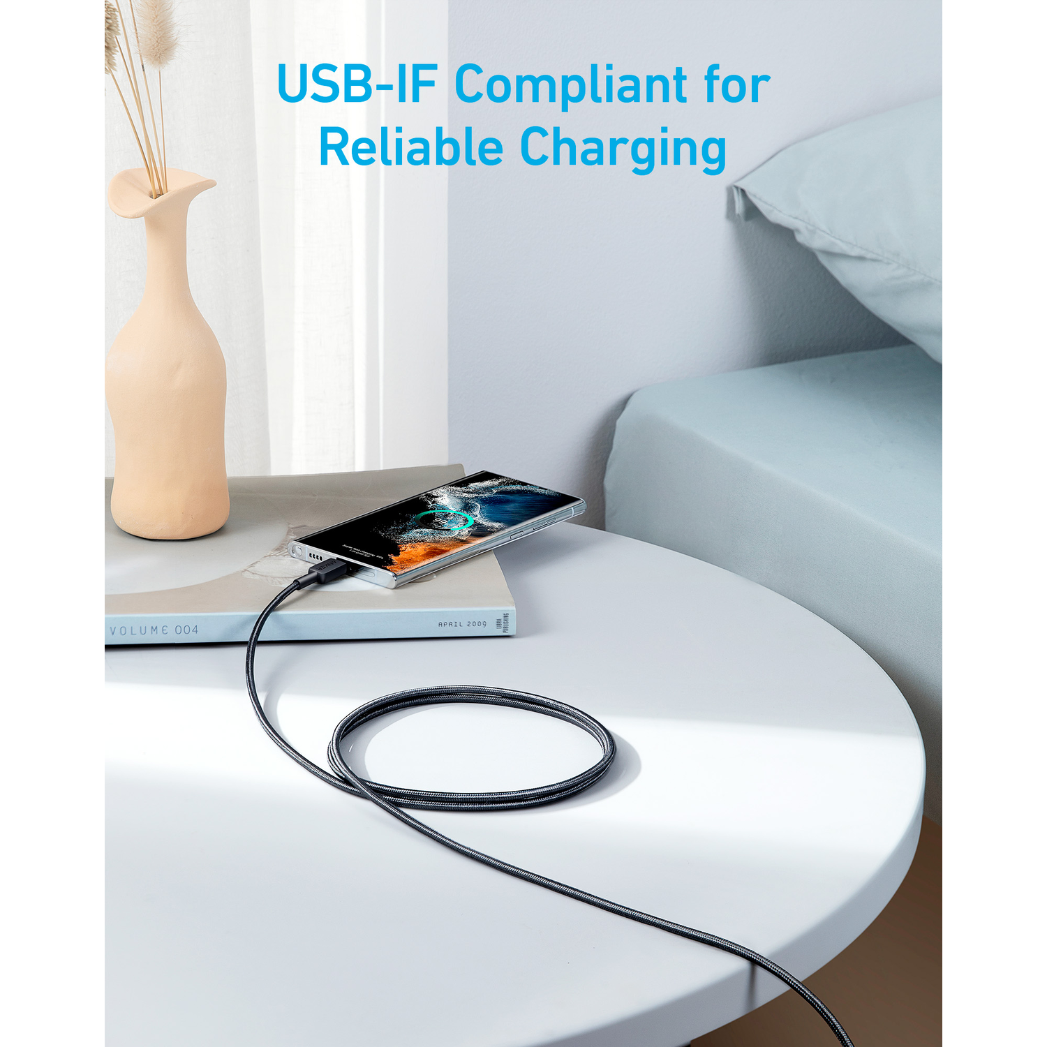 A phone charging on a table with a cable, next to a vase and a book, near a bed.