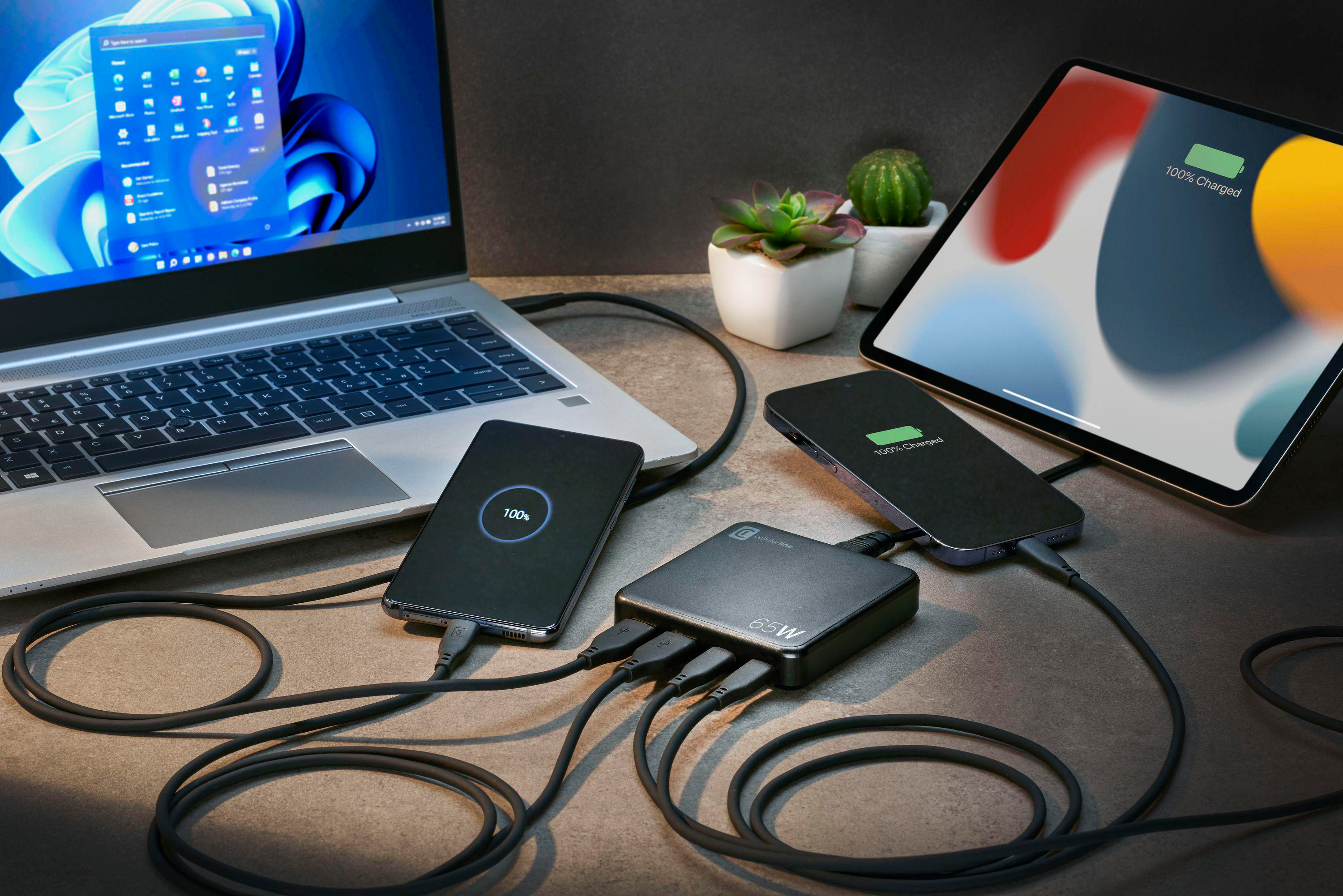 Mehrere Geräte laden: Laptop, Tablet, Telefon und Ladegerät. Kabel sind auf dem Schreibtisch angeschlossen.