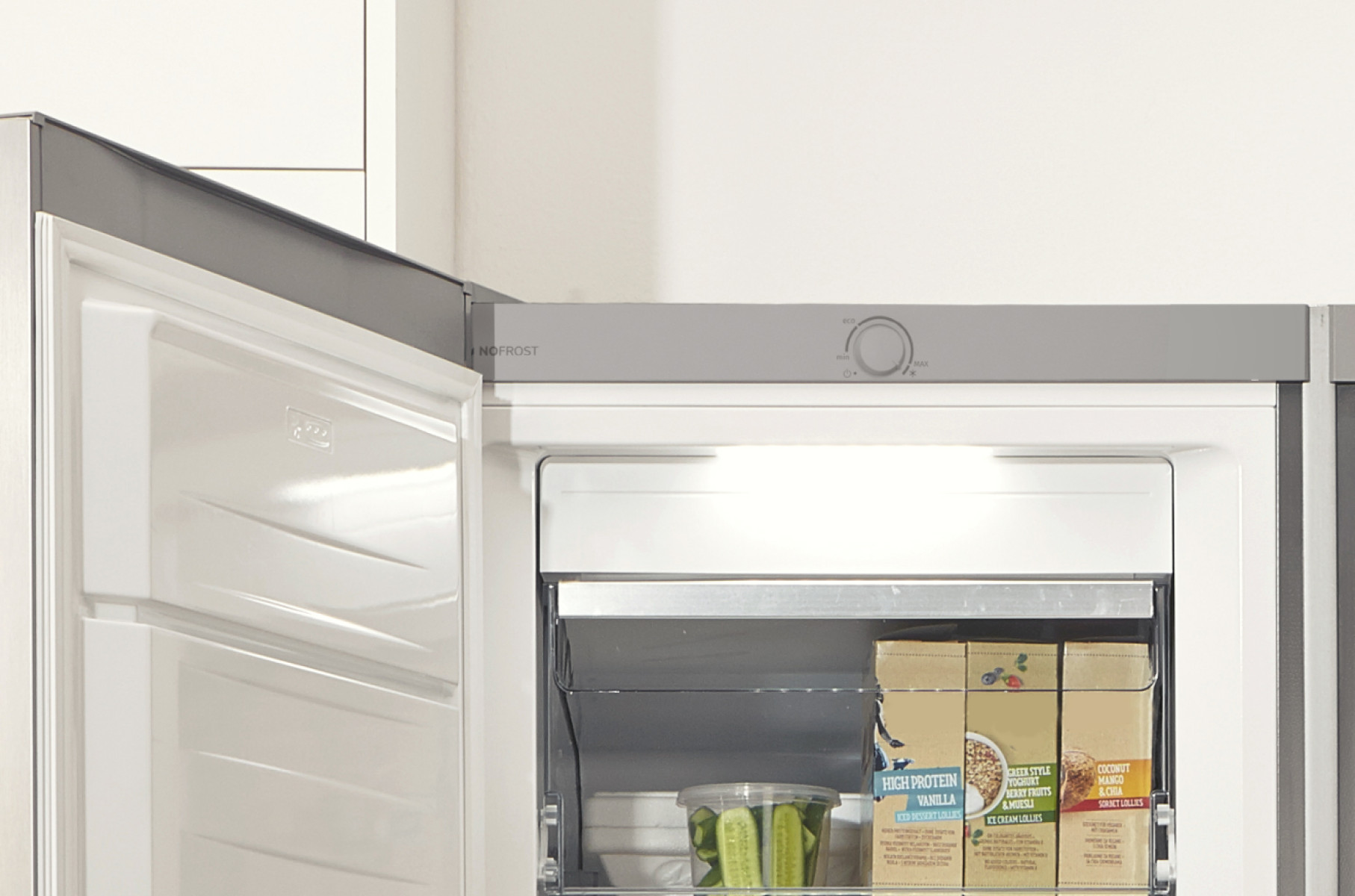 Otwarta lodówka z jedzeniem w środku. Widoczne pudełka z jedzeniem, warzywa i pokrętło.