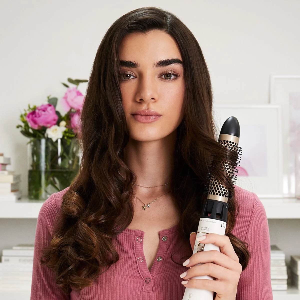 Una donna tiene una spazzola per lo styling dei capelli. Ha lunghi capelli castani e indossa un top rosa.