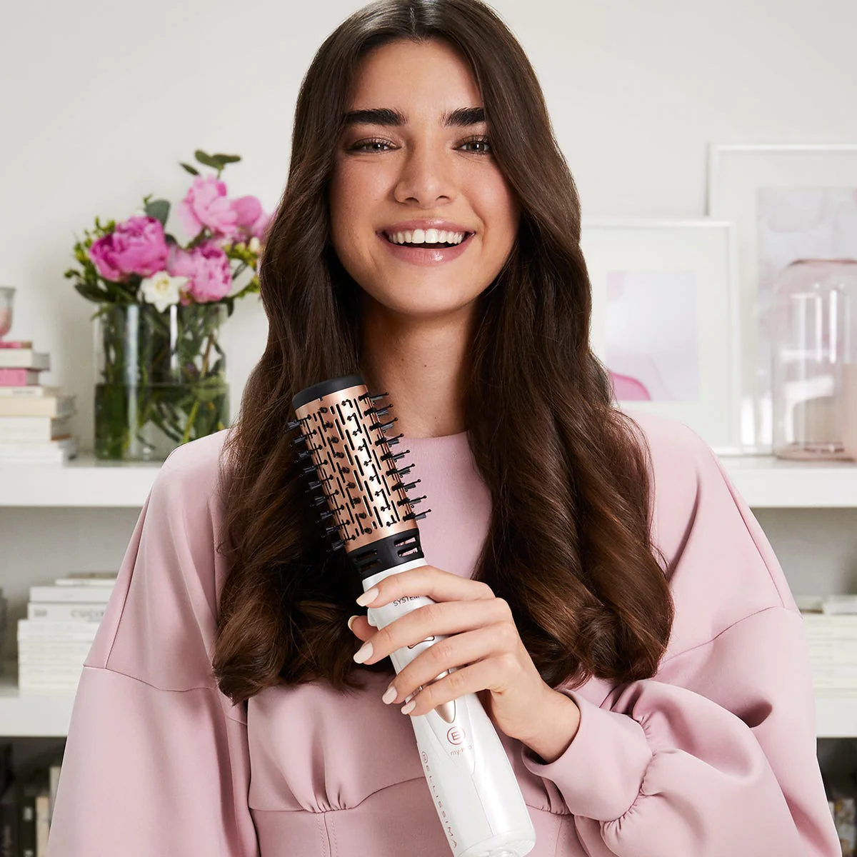 Donna sorridente, che tiene una spazzola per capelli. Maglione rosa, fiori sullo sfondo.