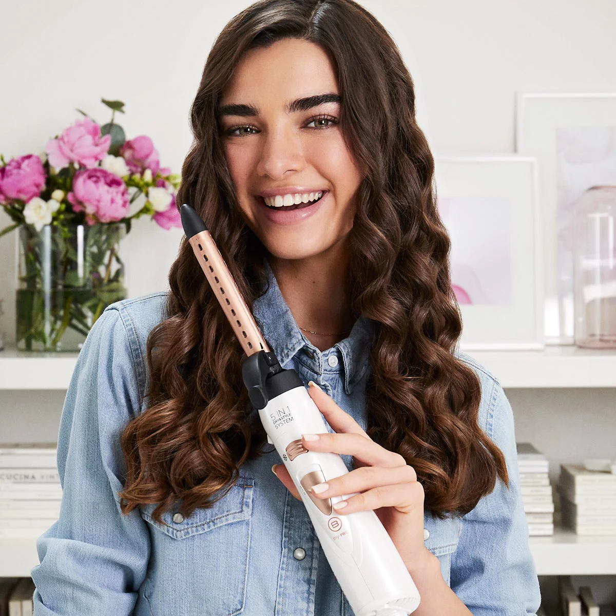Donna sorride, tiene arricciacapelli. Capelli castani e camicia jeans visibili. Fiori rosa sullo sfondo.