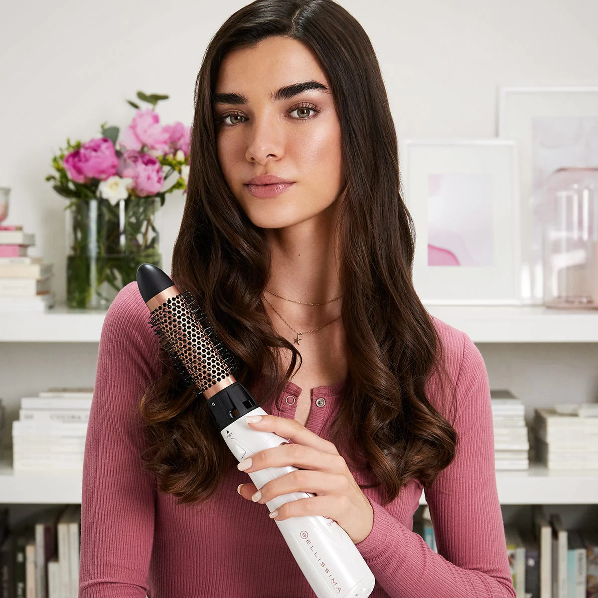 Donna che tiene uno strumento per lo styling dei capelli. Lunghi capelli castani, camicia rosa e sfondo di una libreria.