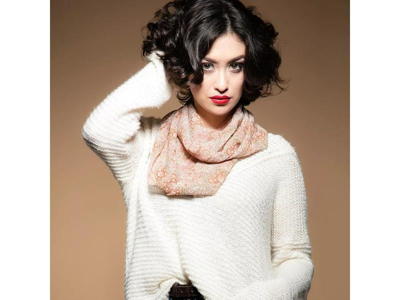 Frau mit dunklem, lockigem Haar, rotem Lippenstift, trägt einen weißen Pullover und einen Schal.
