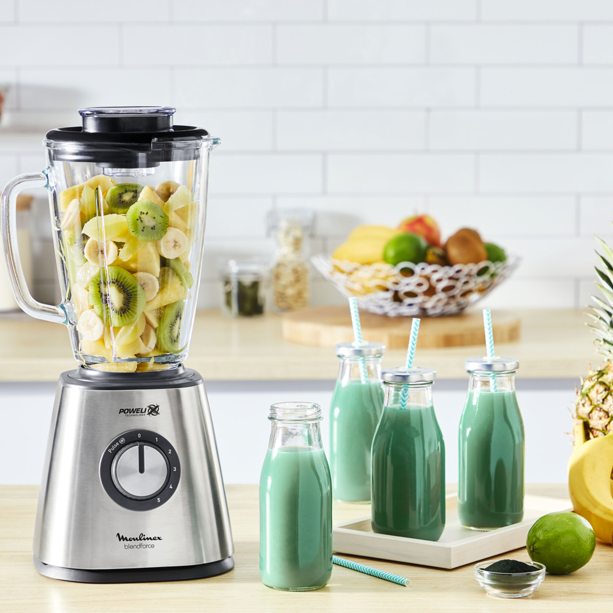 Licuadora con fruta, botellas de zumo y frutero. Piña y lima a la derecha.