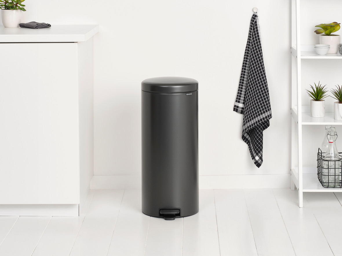 Dunkelgrauer Mülleimer. Weiße Wände und Boden. Schwarz-weißes Handtuch an der Wand. Weißes Regal.