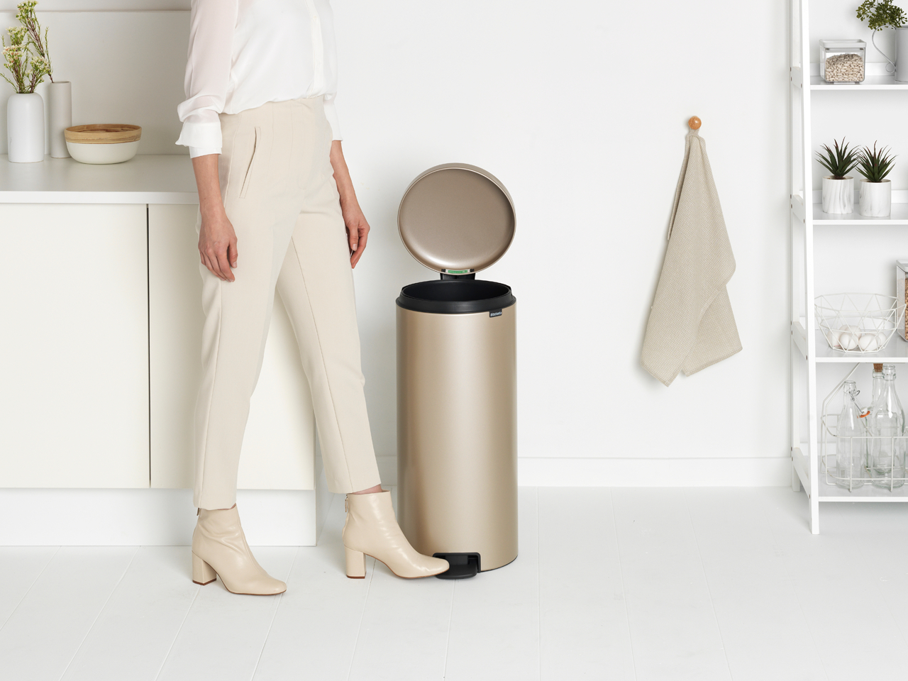 Eine Frau in beiger Hose und Stiefeln steht in einer hellen Küche neben einem goldenen Mülleimer.