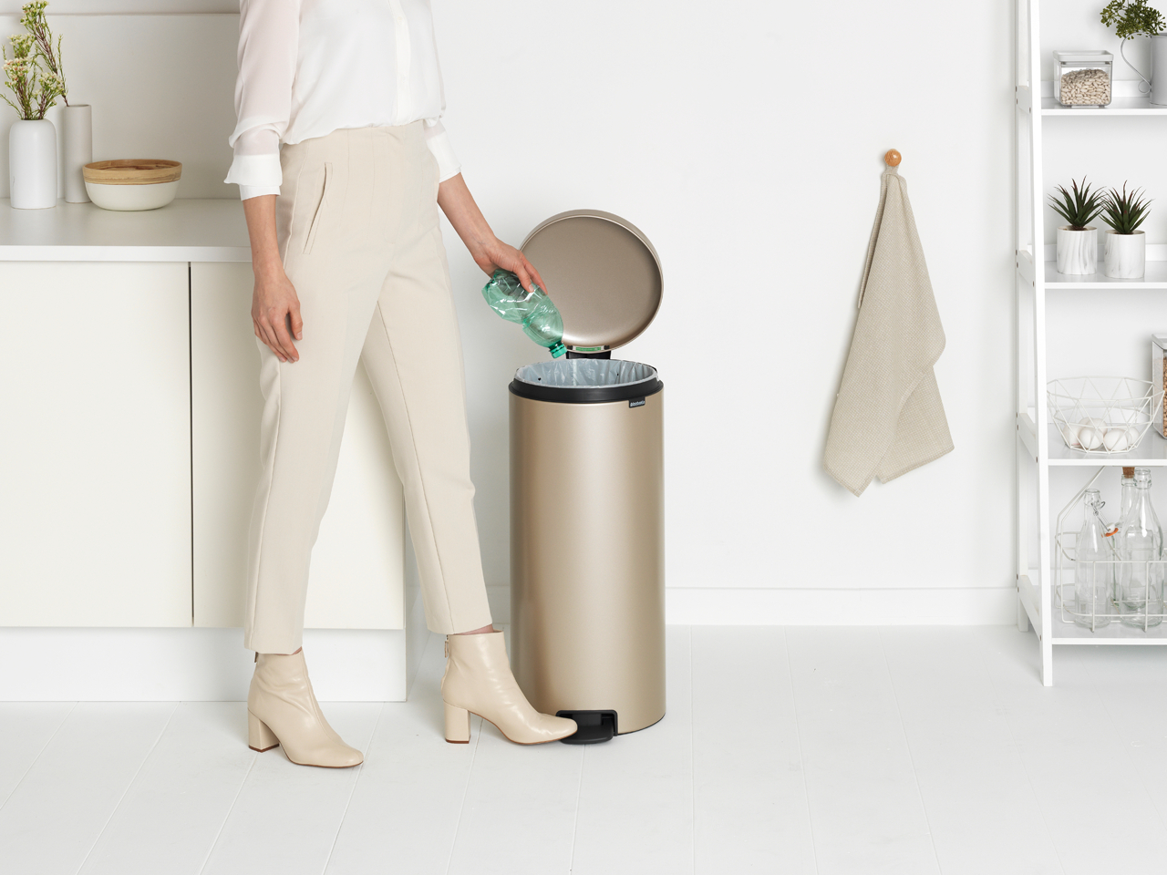 Frau wirft Plastikflasche in einen goldenen Mülleimer. Weiße Wand, Regal und Handtuch.