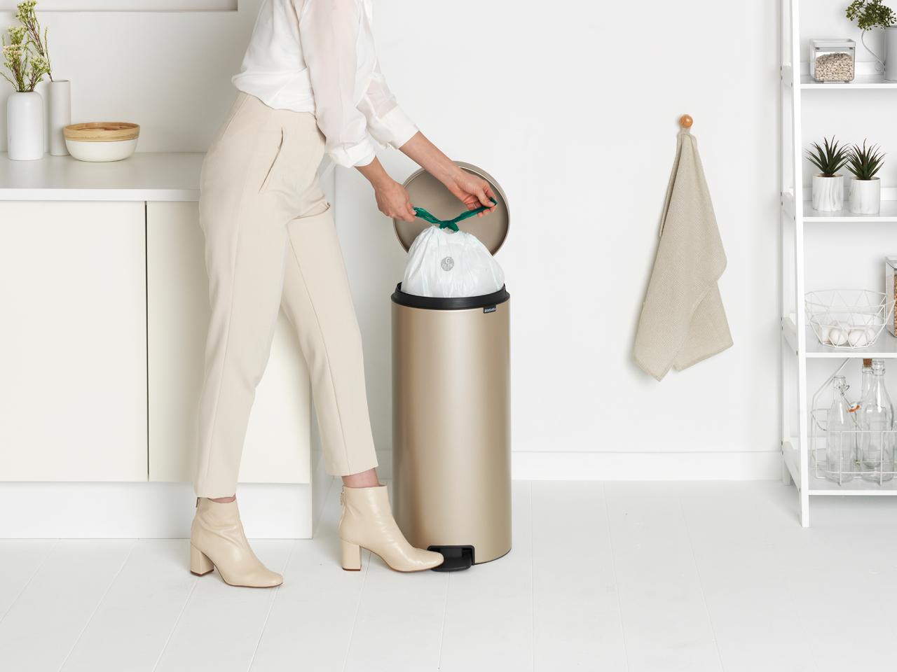Frau, die einen weißen Müllsack in einen goldenen Mülleimer gibt. Küchenumgebung mit weißen Schränken und einem Regal.