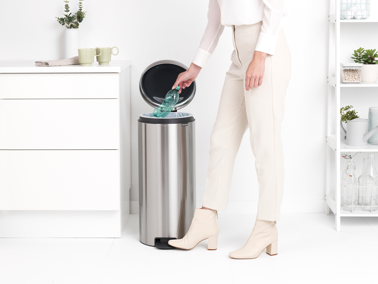Eine Frau wirft eine Flasche in einen Mülleimer aus Edelstahl. Weiße Schränke und Regale sind im Hintergrund.