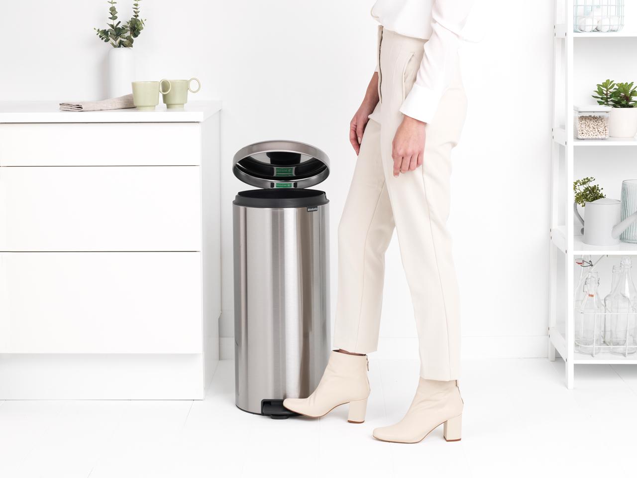 Frau neben einem silbernen Mülleimer, trägt beige Kleidung, in einem weißen Raum.