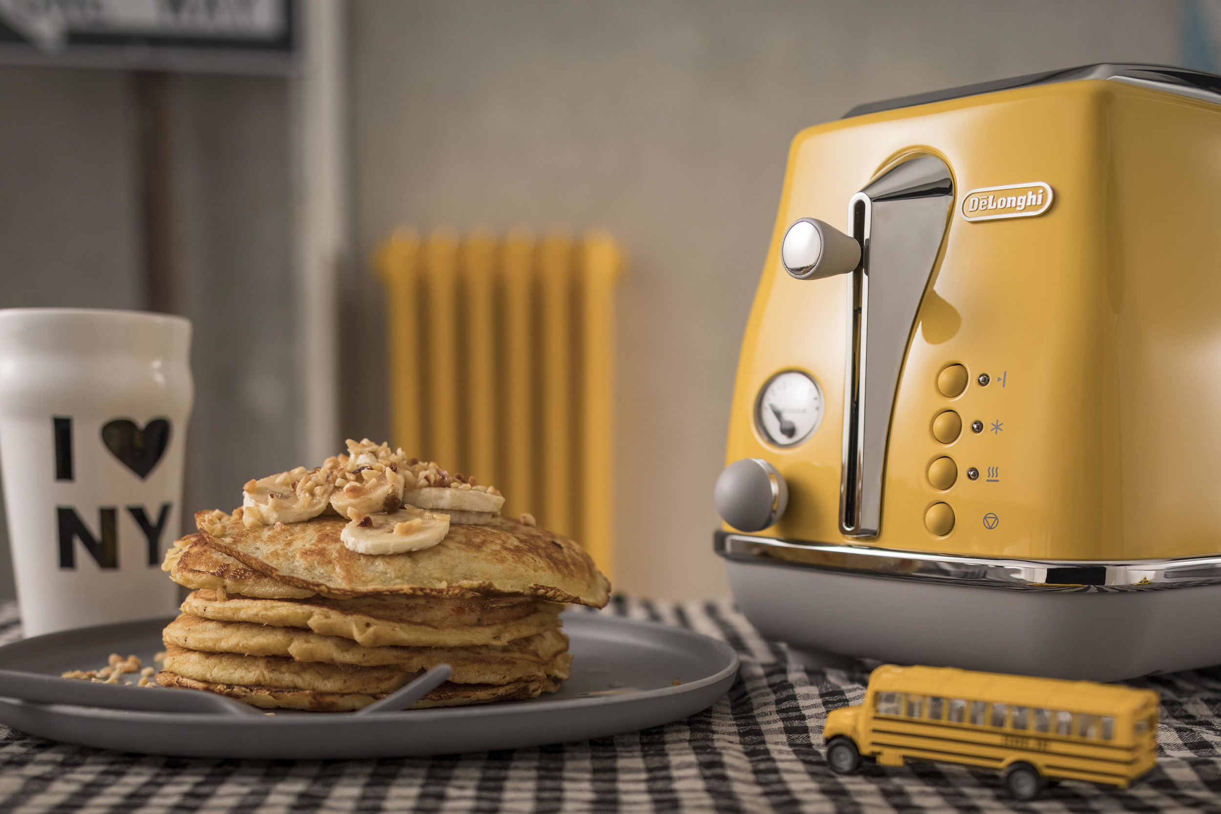 Stos naleśników z bananami, żółty toster i kubek NY na stole.