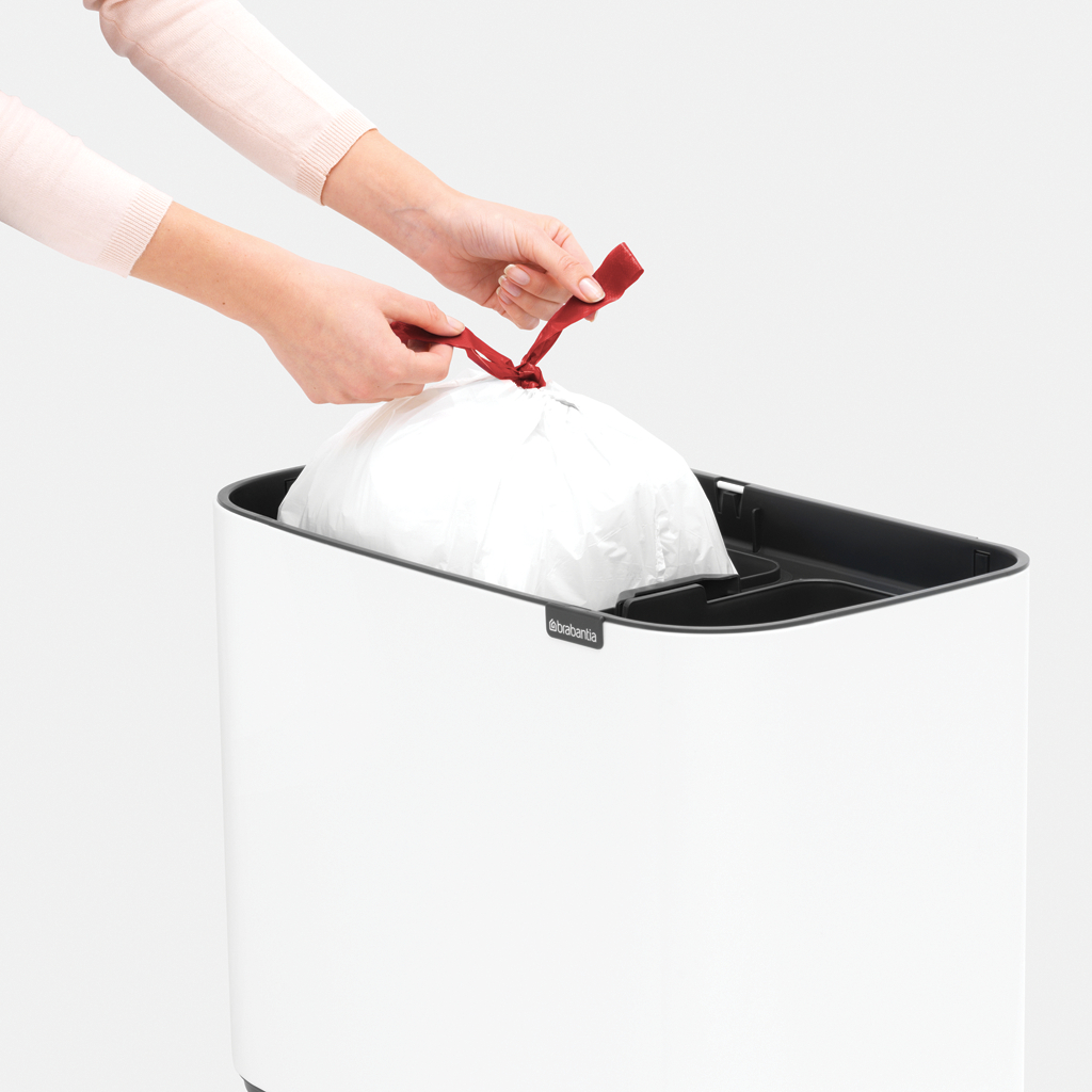 Hände binden ein rotes Band an einem weißen Müllsack. Der Sack ist in einem weißen Mülleimer.
