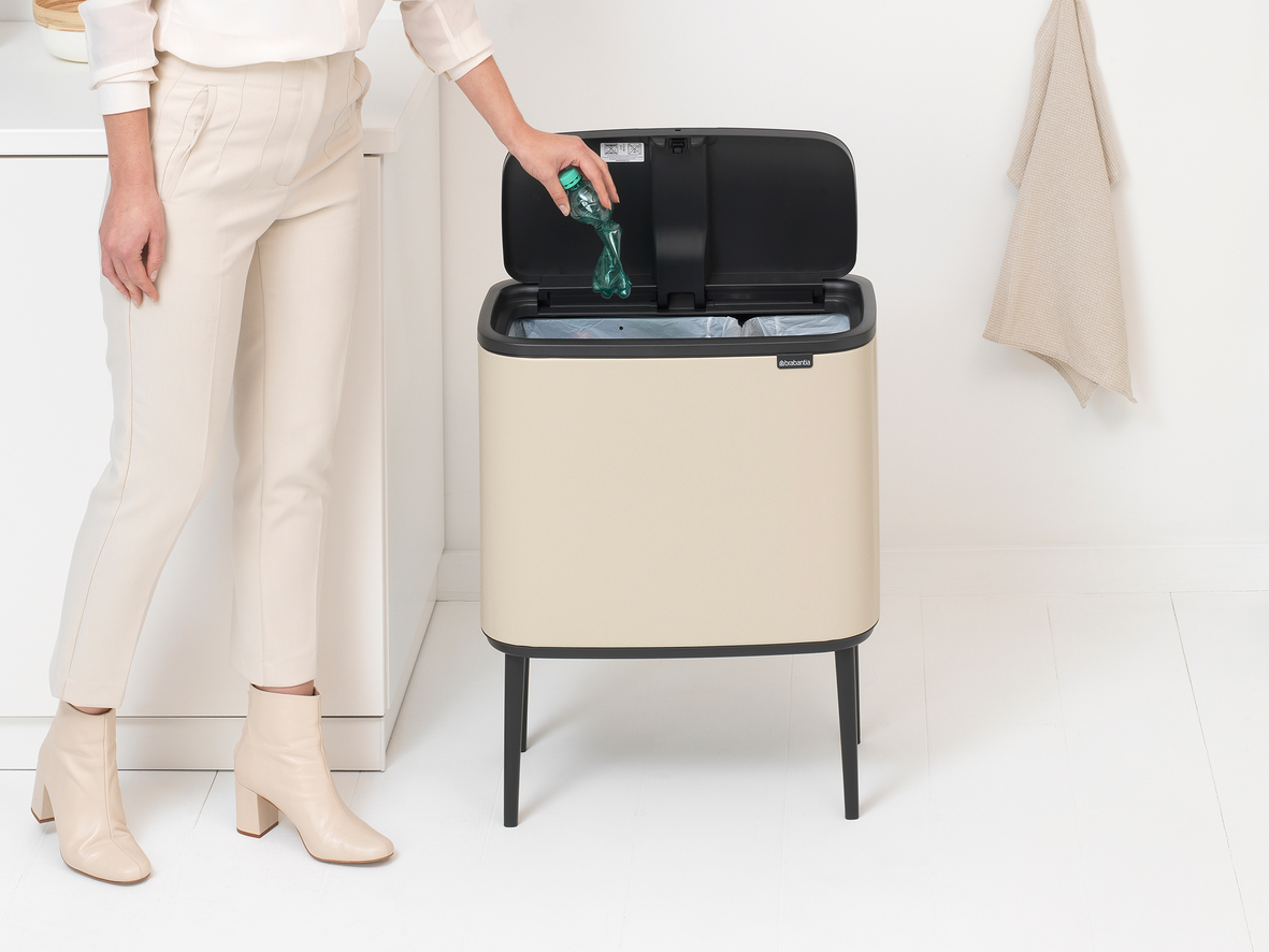 Eine Frau wirft eine Plastikflasche in einen beigefarbenen Brabantia-Mülleimer mit schwarzem Deckel, in der Nähe eines Handtuchs.