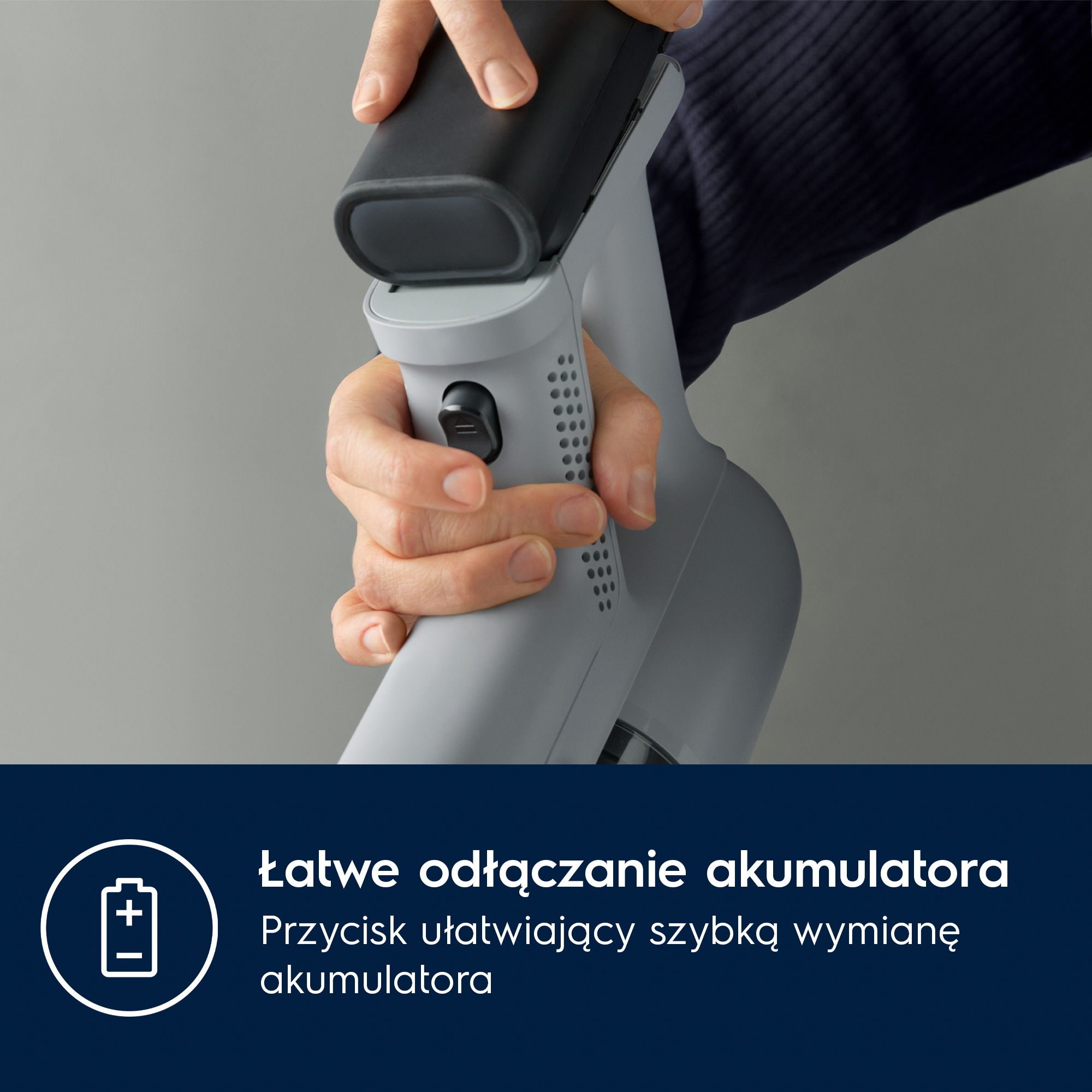 Ręka wyjmuje baterię z szarego odkurzacza.