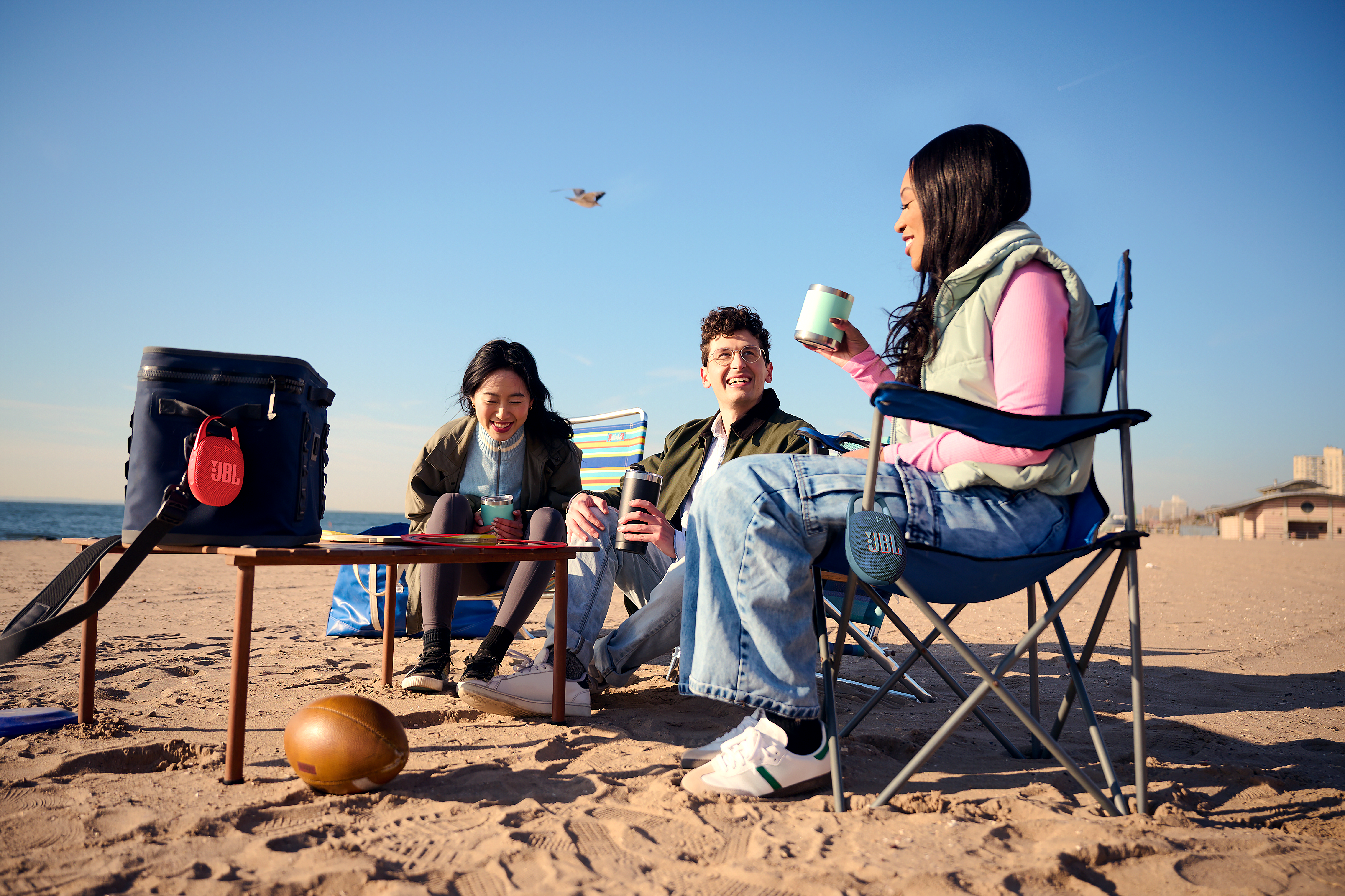 Drei Personen sitzen am Strand mit JBL-Lautsprechern und Kühler.