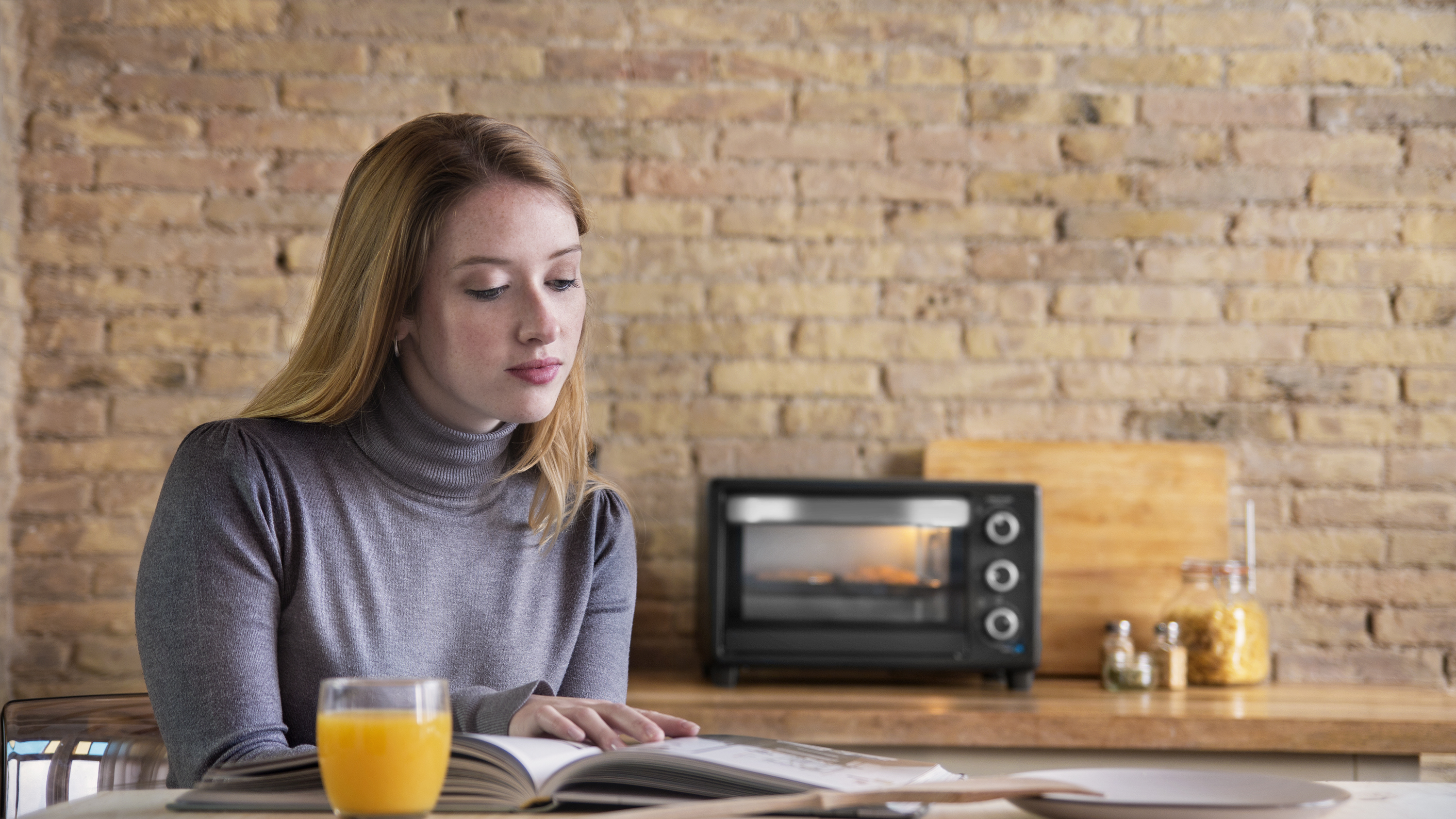 Una mujer lee un libro en una mesa con un horno tostador y zumo de naranja.