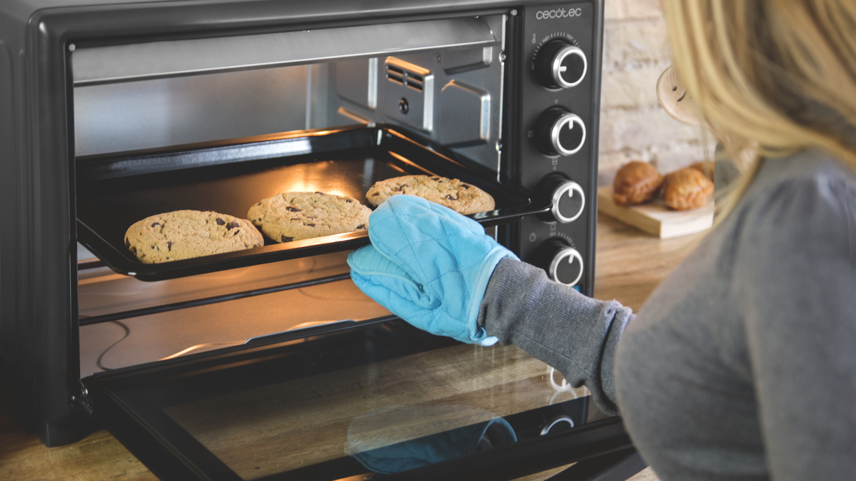 Mujer sacando galletas de un horno negro con un guante azul. Galletas en la bandeja.