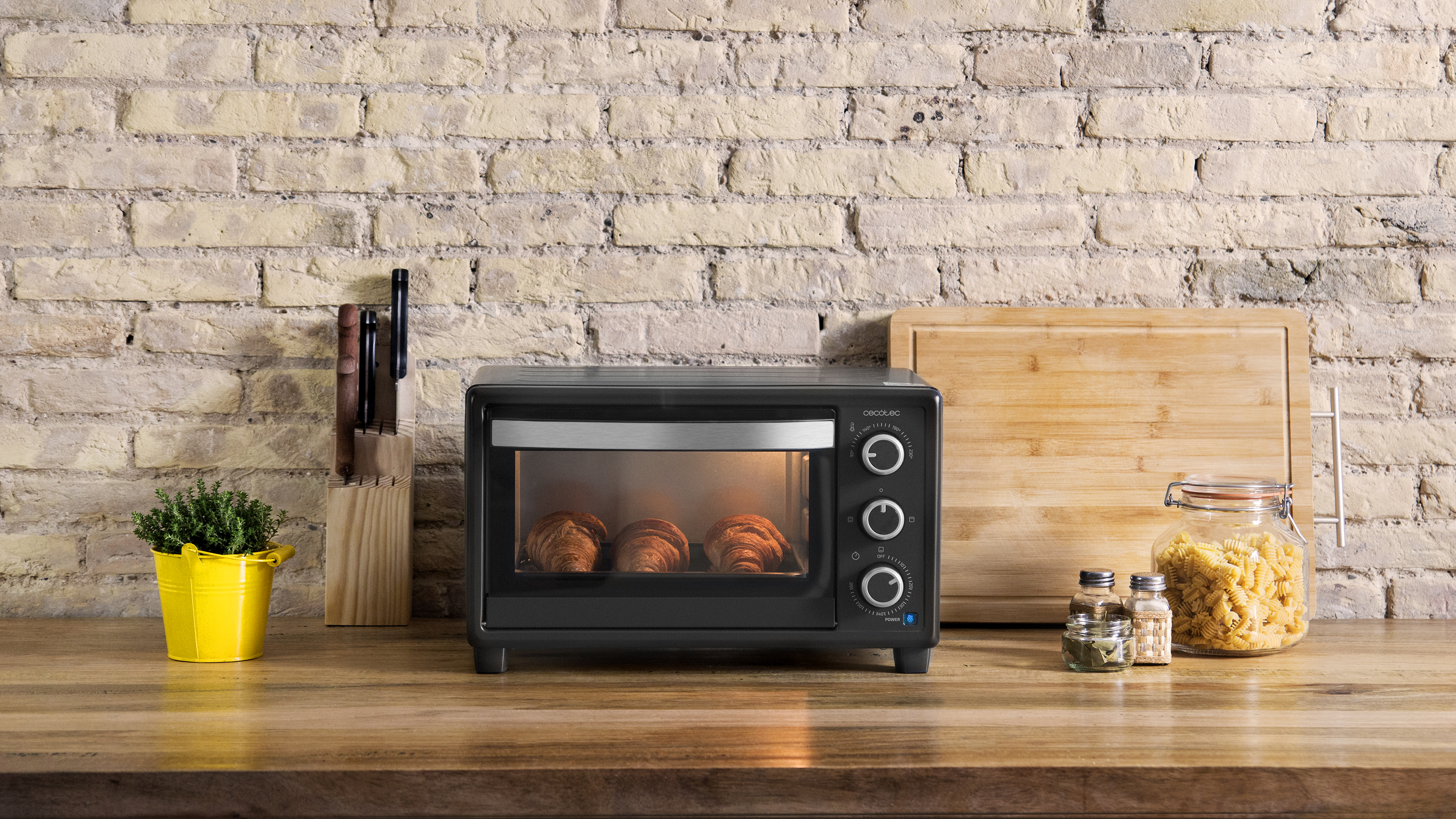 Horno negro con cruasanes dentro. Encimera de madera. Fondo de pared de ladrillo. Accesorios como cuchillo, hierbas, especias.