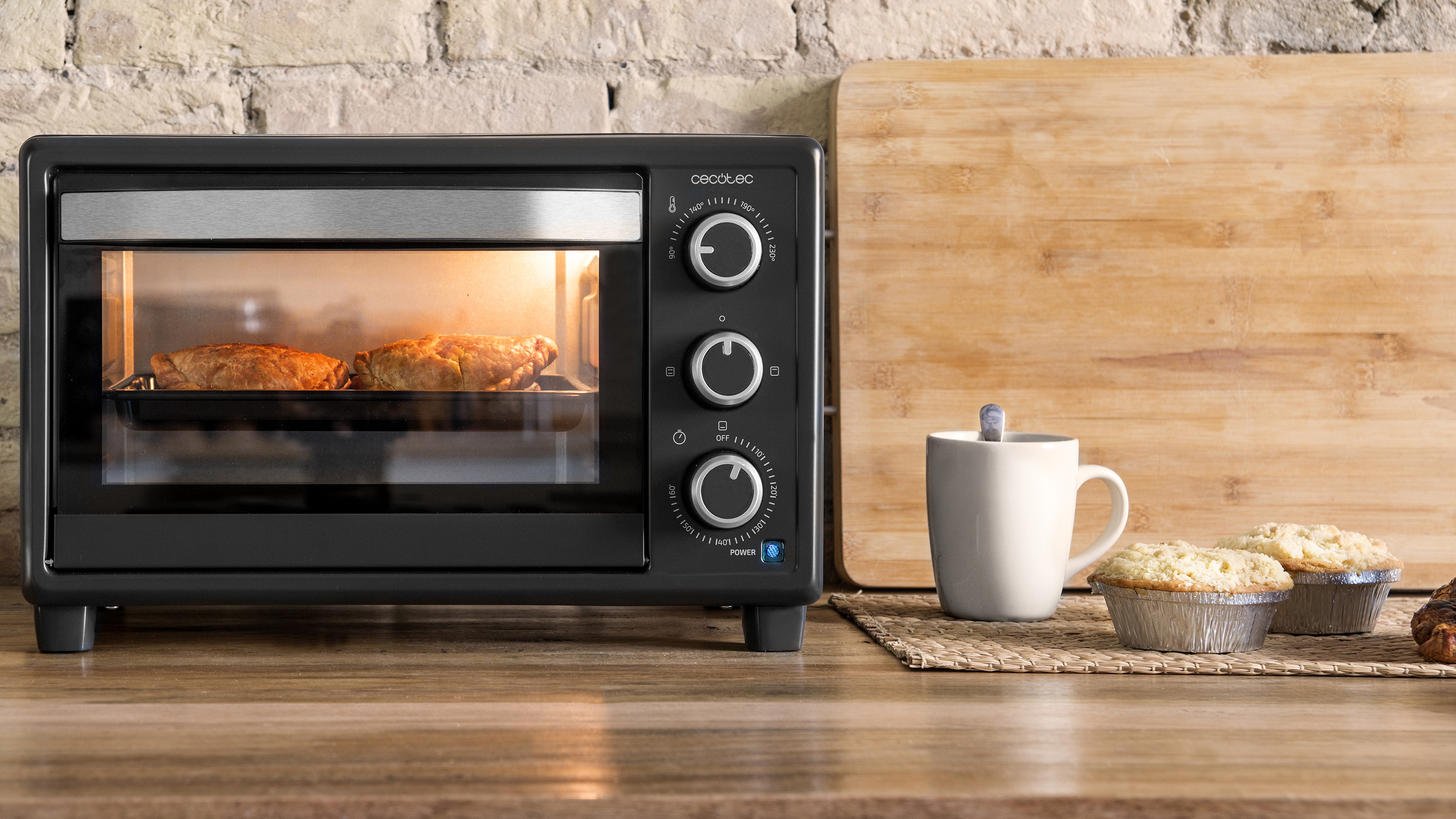 Mini horno negro sobre superficie de madera. Dos platos horneados dentro. Taza blanca y magdalenas cerca.