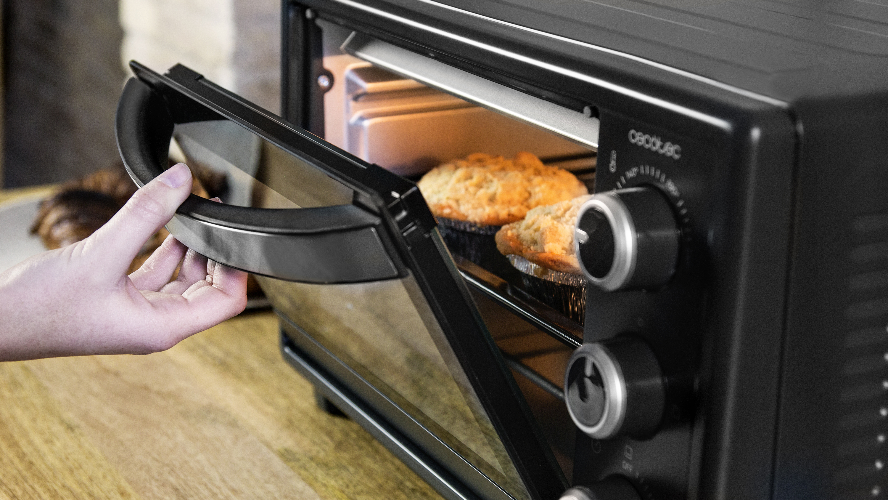 Una mano abriendo un horno negro con magdalenas dentro.