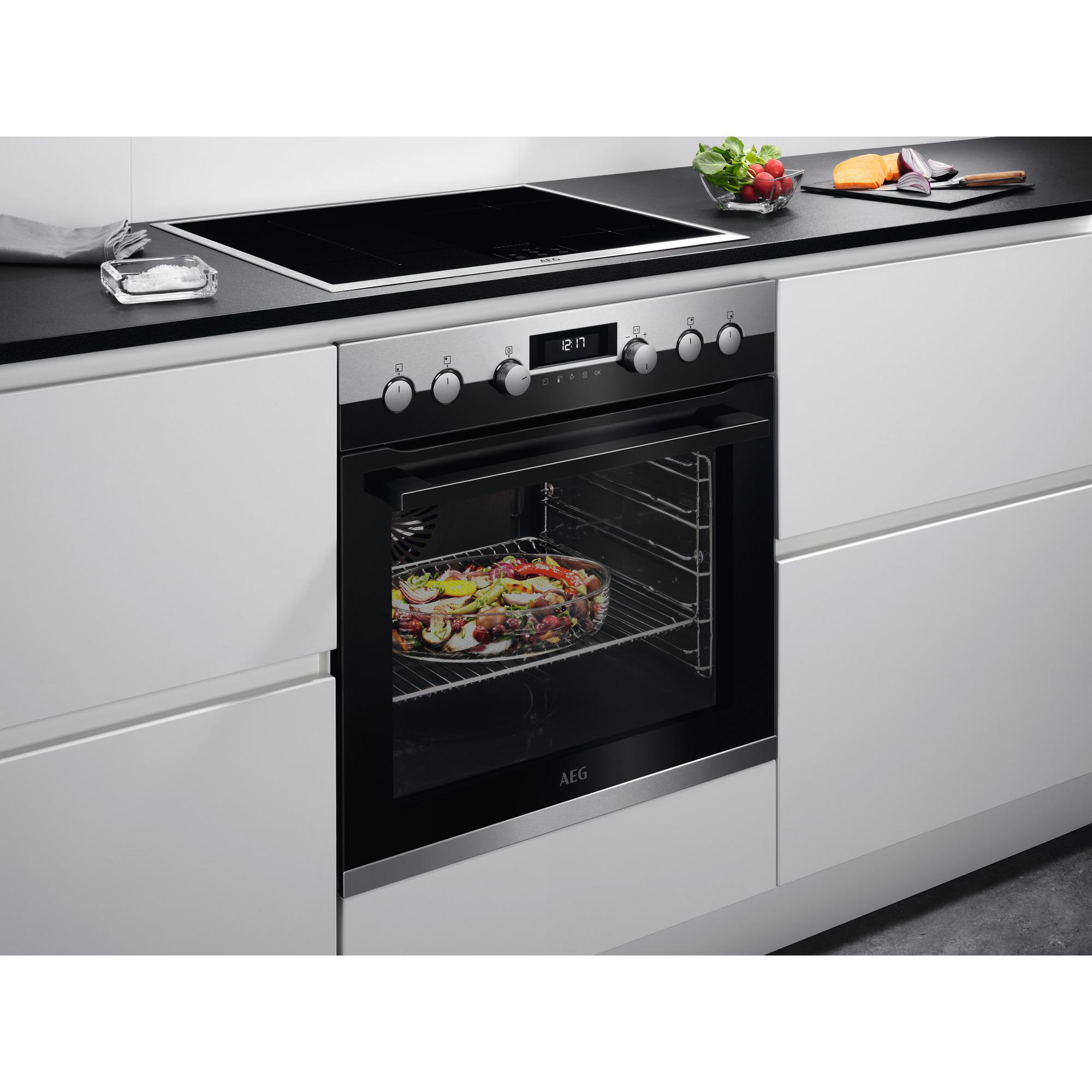 Küche mit Backofen und Induktionskochfeld. Ein Tablett mit Essen befindet sich im Ofen. Gemüse auf der Theke.
