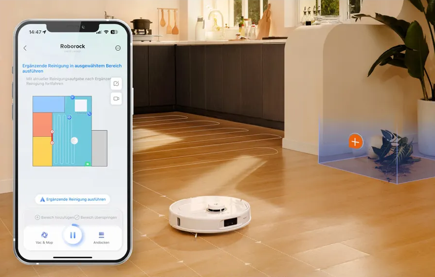 Smartphone con app, accanto a un robot aspirapolvere. Cucina e pavimento in legno sullo sfondo.