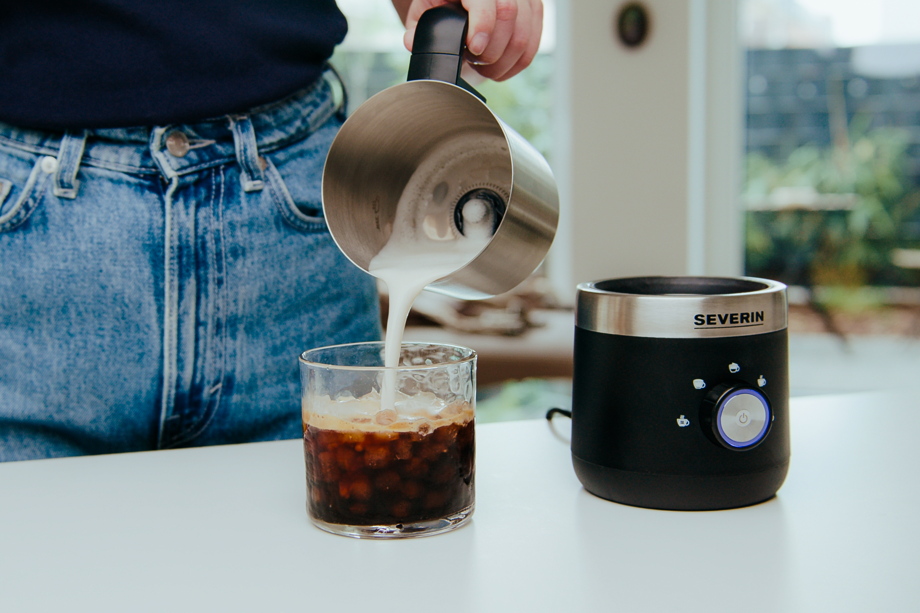 Person gießt Milch aus silberner Kanne in Glas mit Eis und Kaffee, auf weißem Tisch.