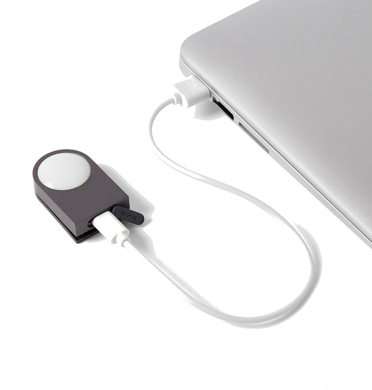 Szare urządzenie z białym światłem jest połączone z laptopem za pomocą białego kabla. Urządzenie jest również ładowane przez USB.