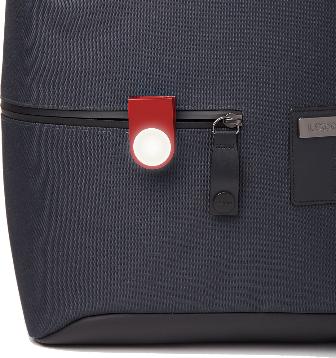 Zbliżenie na ciemnoszarą torbę z czerwoną lampką na klips. Widoczny zamek błyskawiczny i skórzana metka. Marka 'LEXON' na torbie.