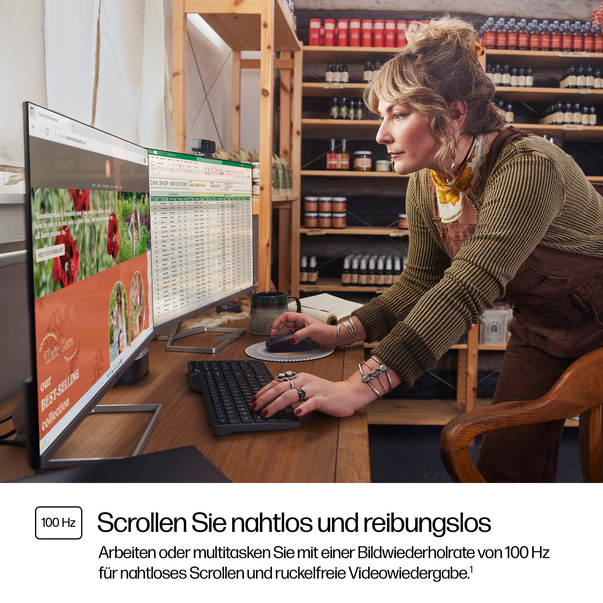 Eine Frau arbeitet an einem Schreibtisch mit einem Monitor, der eine Website und eine Tabelle anzeigt. Regale mit Produkten sind im Hintergrund.