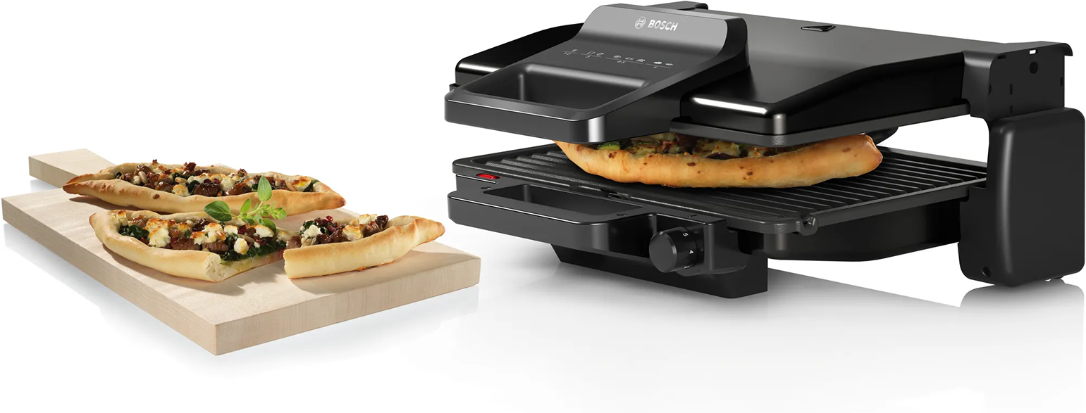 Fekete Bosch pizza grill pizzát süt. Két pizza egy fa deszkán balra.